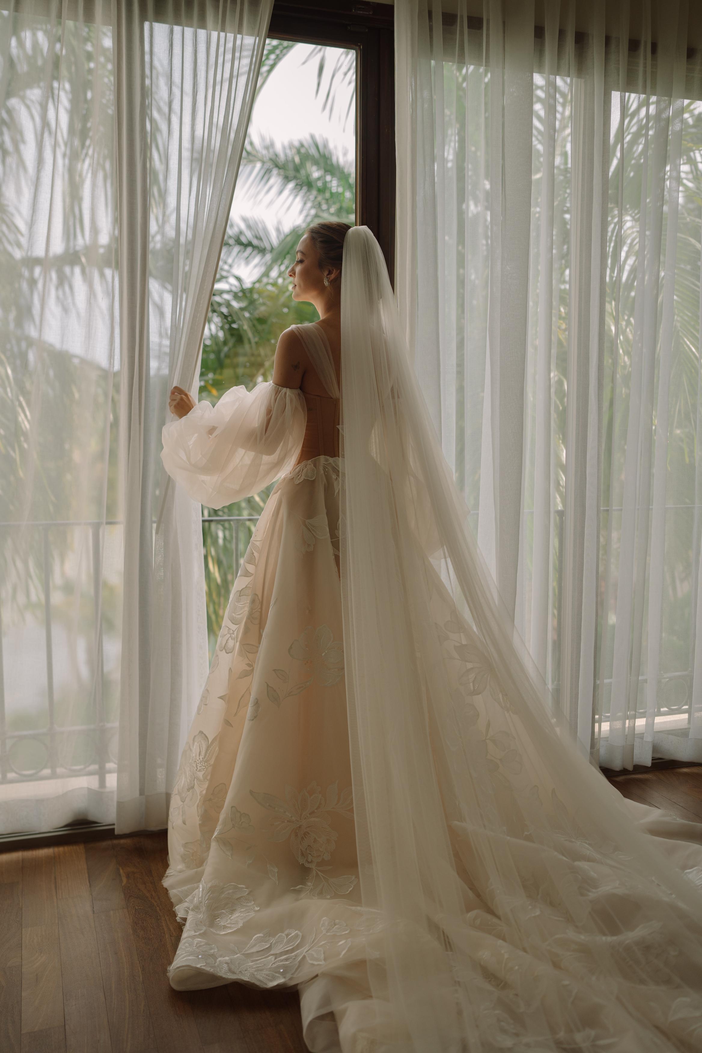 GL Bride Carina wearing the Evelyn wedding gown, with a long train and veil standing in front of a large window with sheer curtains.