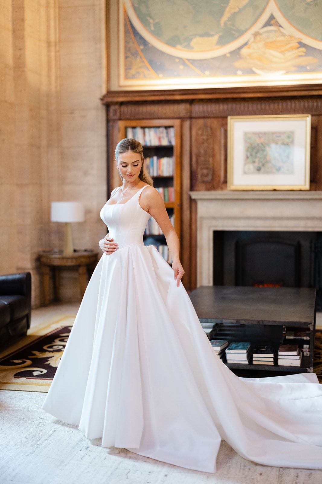GL Bride Eliza in our Dove standing in an elegant room with a fireplace and artwork.