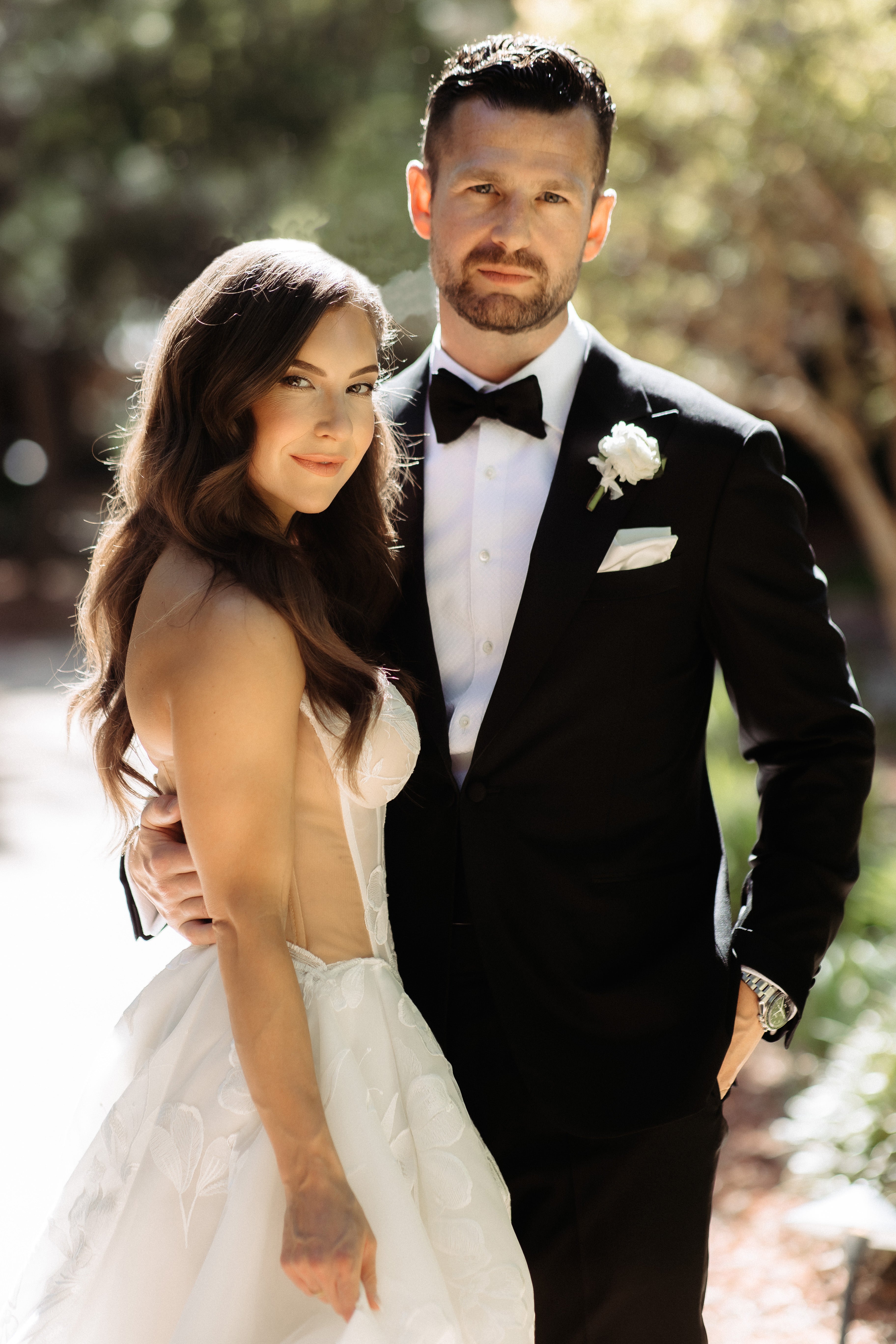 Man in a black tuxedo and woman in our Evelyn wedding dress standing together outdoors.