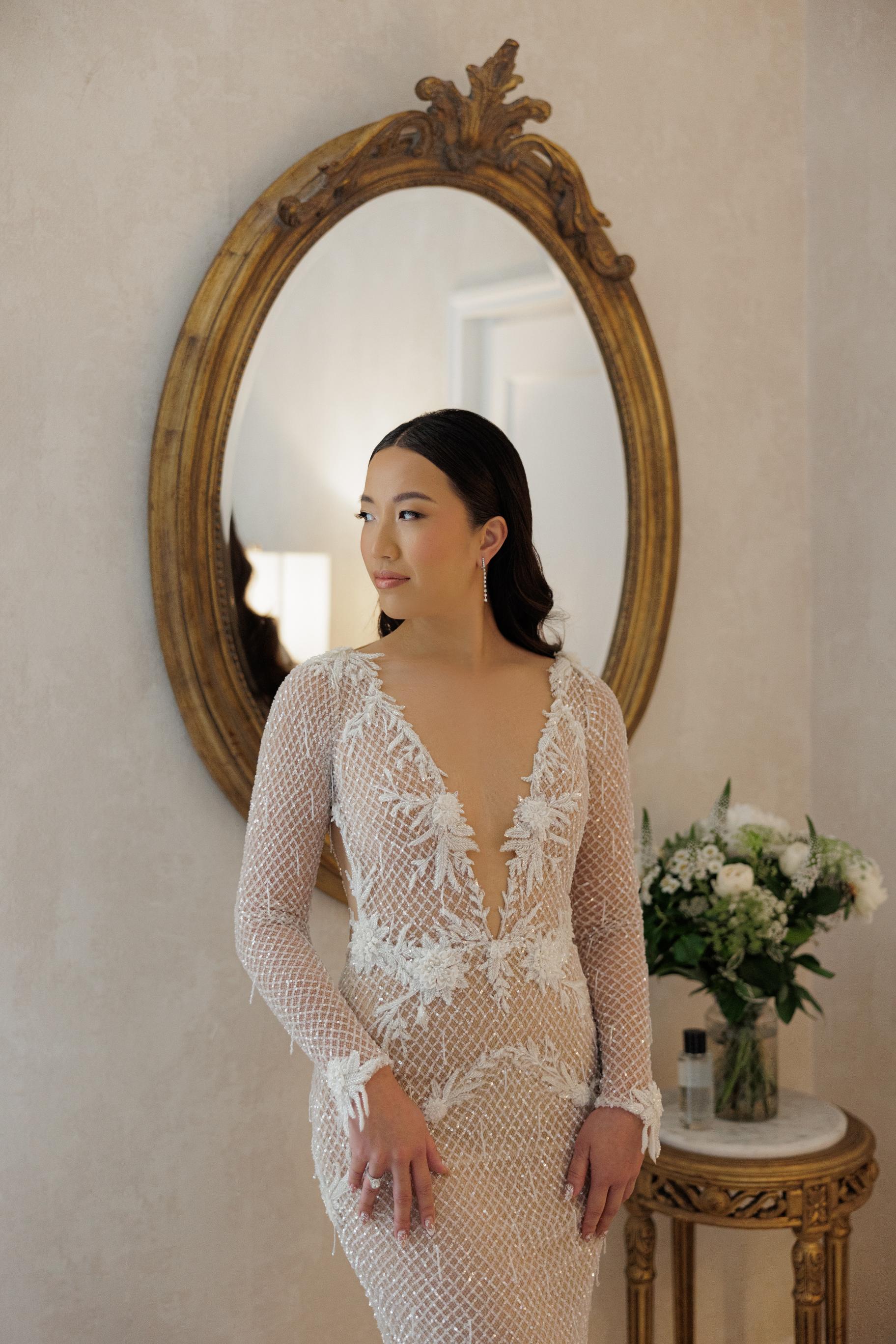 Woman in a lace wedding dress standing in front of an ornate mirror.
