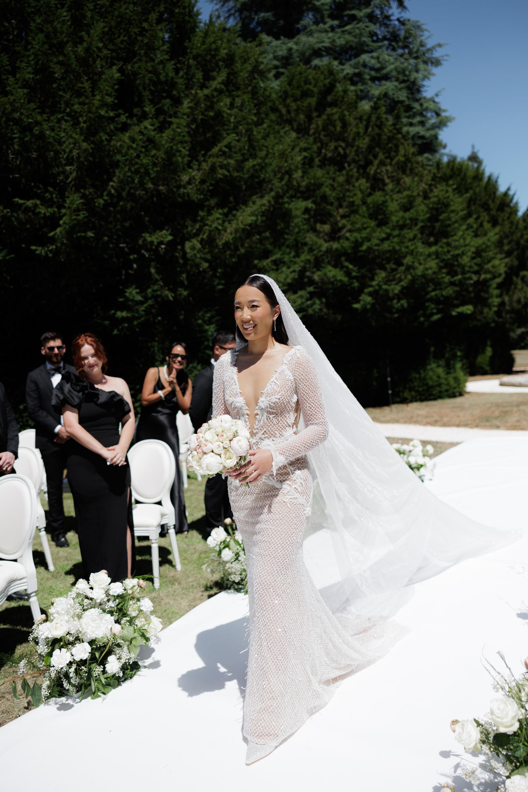 Woman in a wedding dress with a veil walking outdoors with floral decorations.