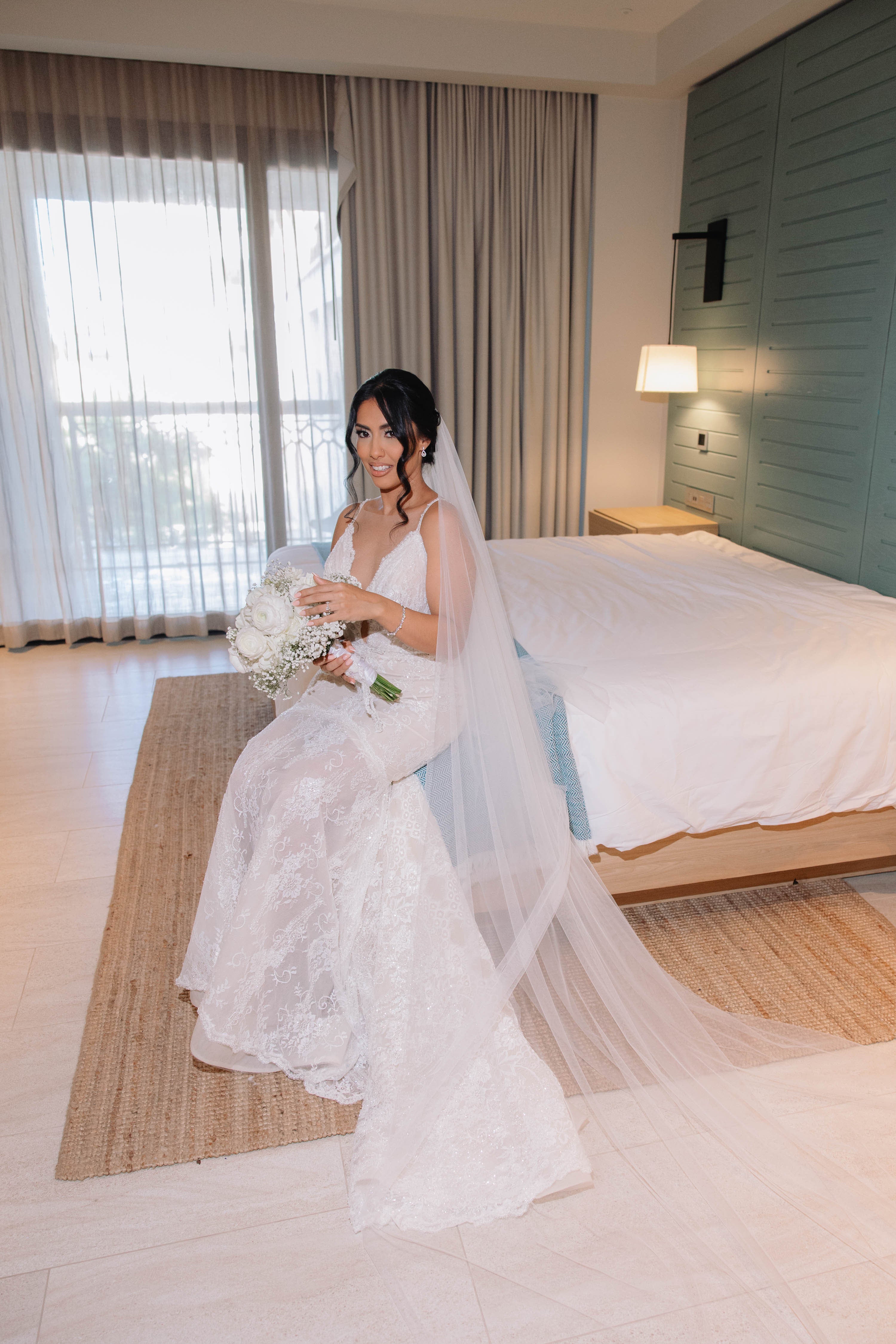 Bride in a white wedding dress with a long veil sitting on a bed in a well-lit room.