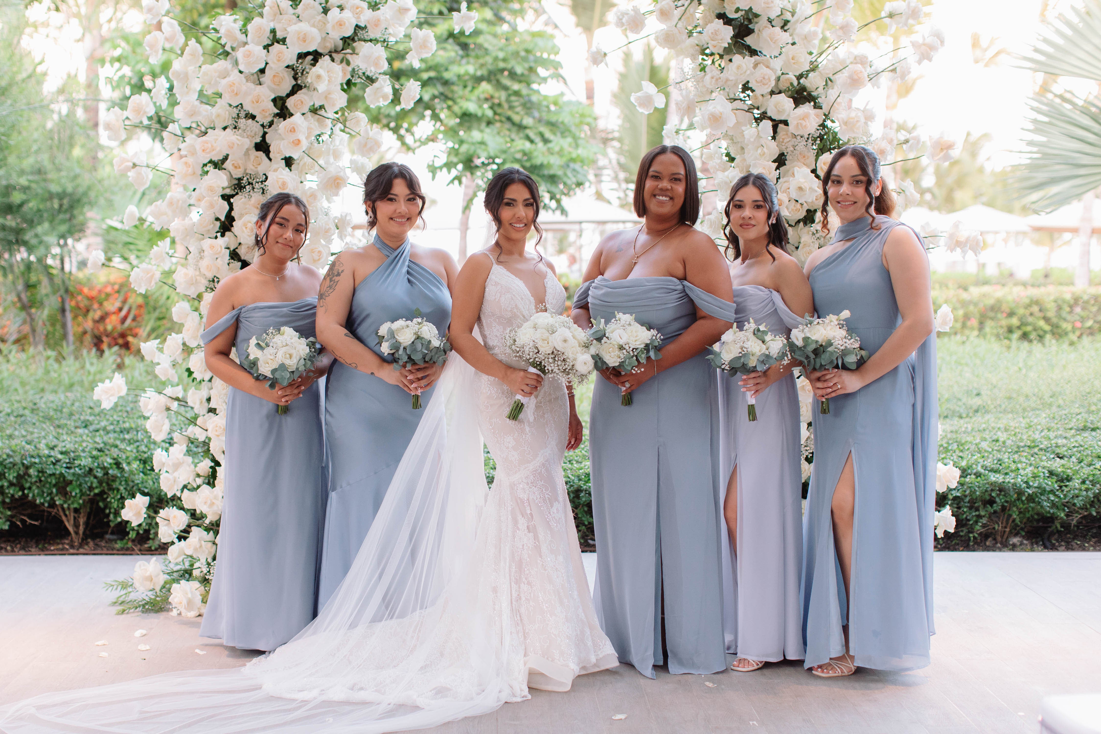 Bride with bridesmaids in light blue dresses standing in front of a floral arch.