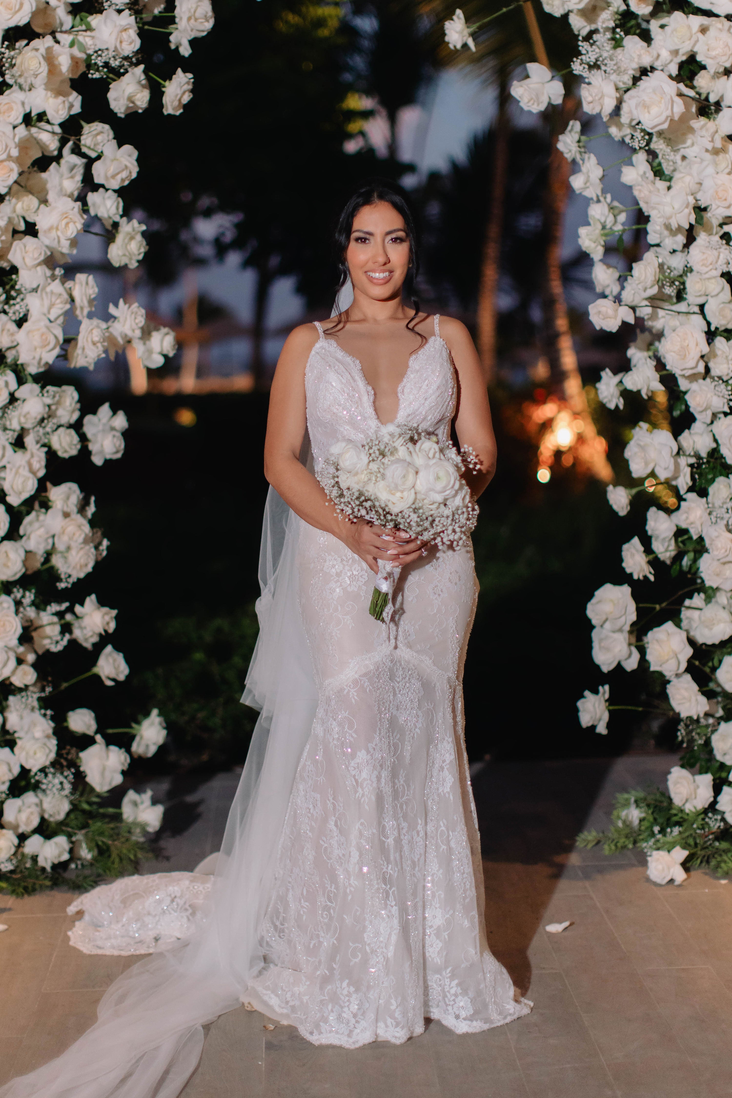 Bride in a white wedding dress holding a bouquet of flowers, standing in front of a floral arch.