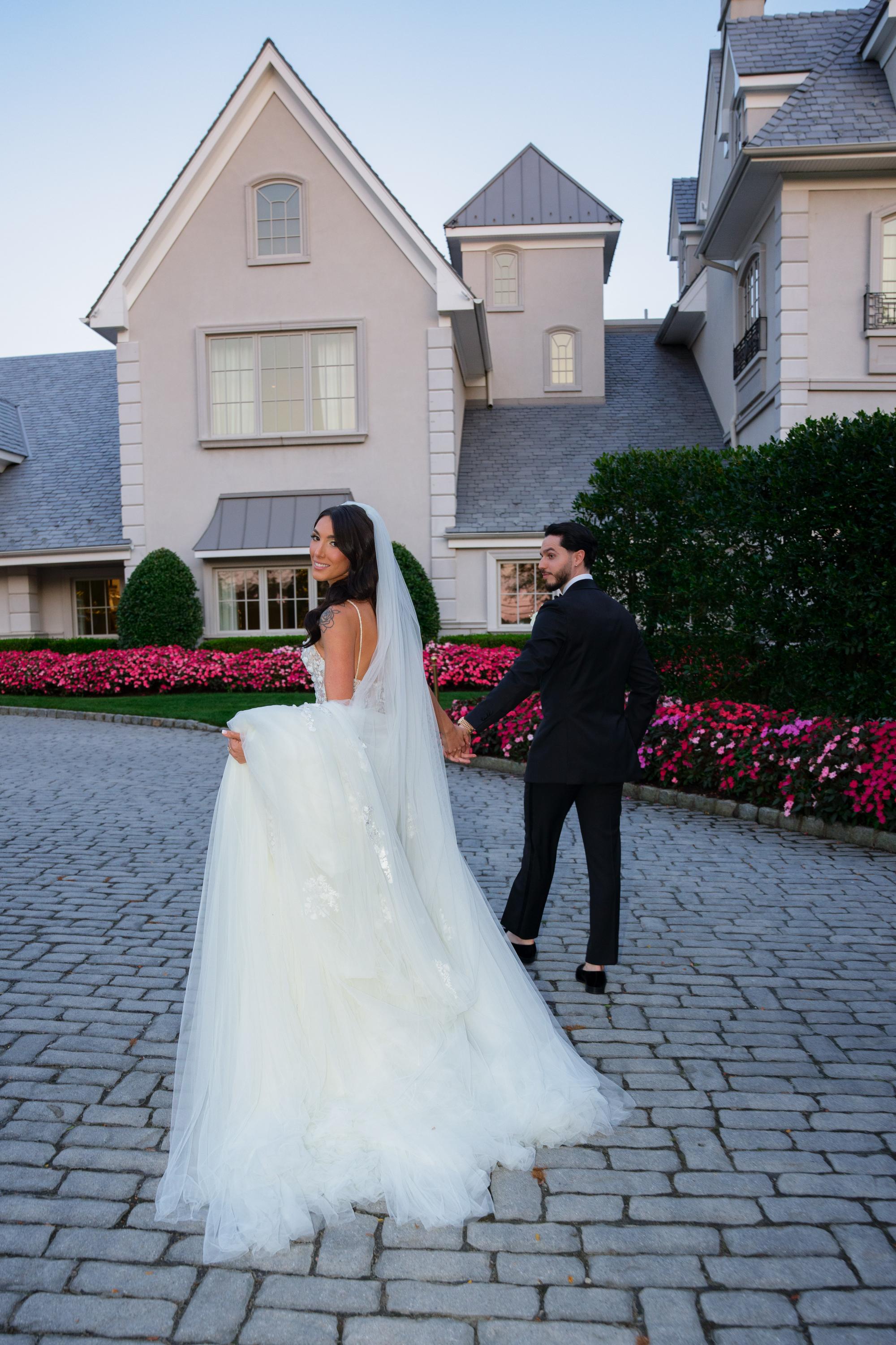 GL Bride Jessica wearing our Catalina gown and her husband in wedding attire standing on a cobblestone driveway in front of a house.