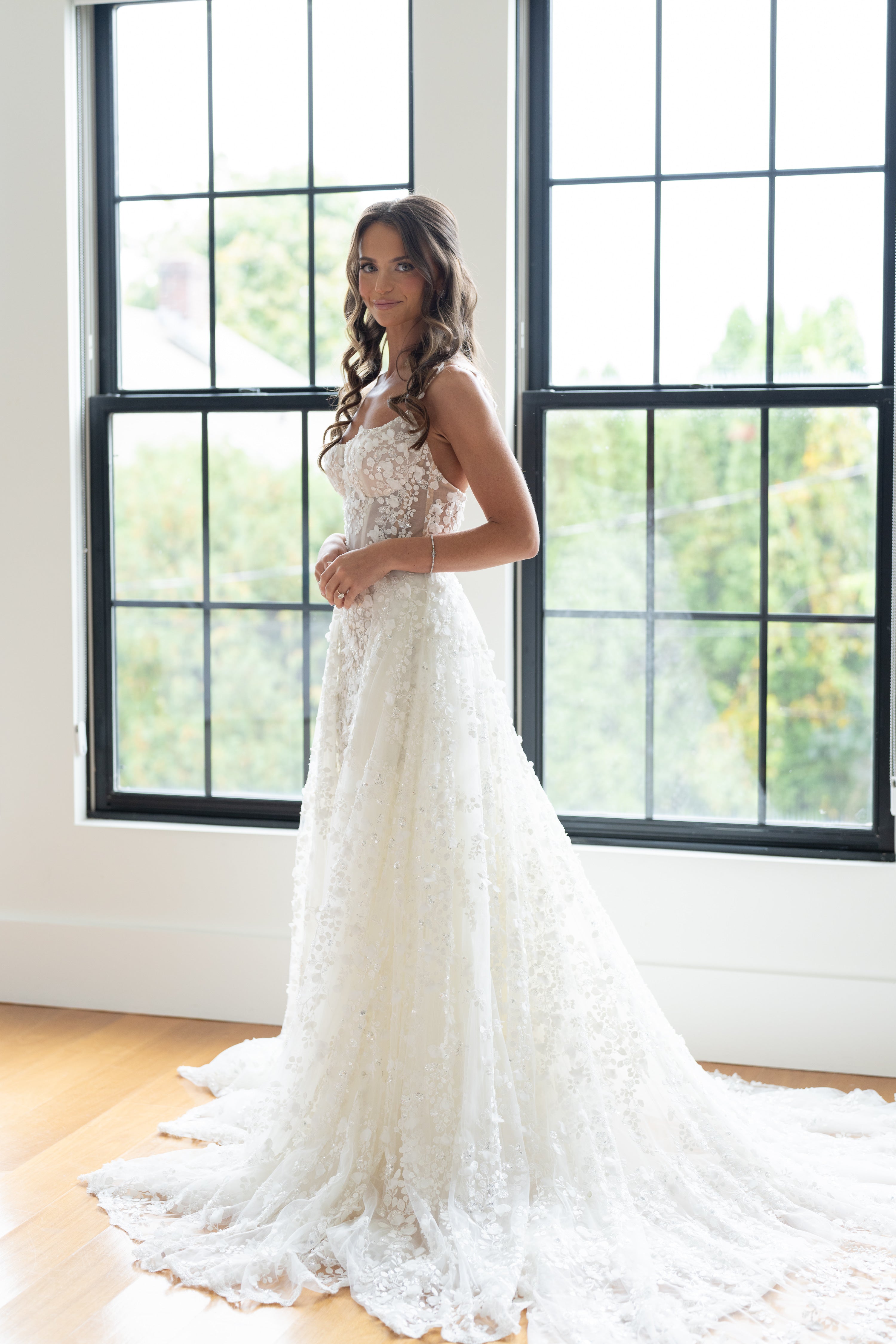 Woman in a white lace wedding dress standing in a room with large windows.