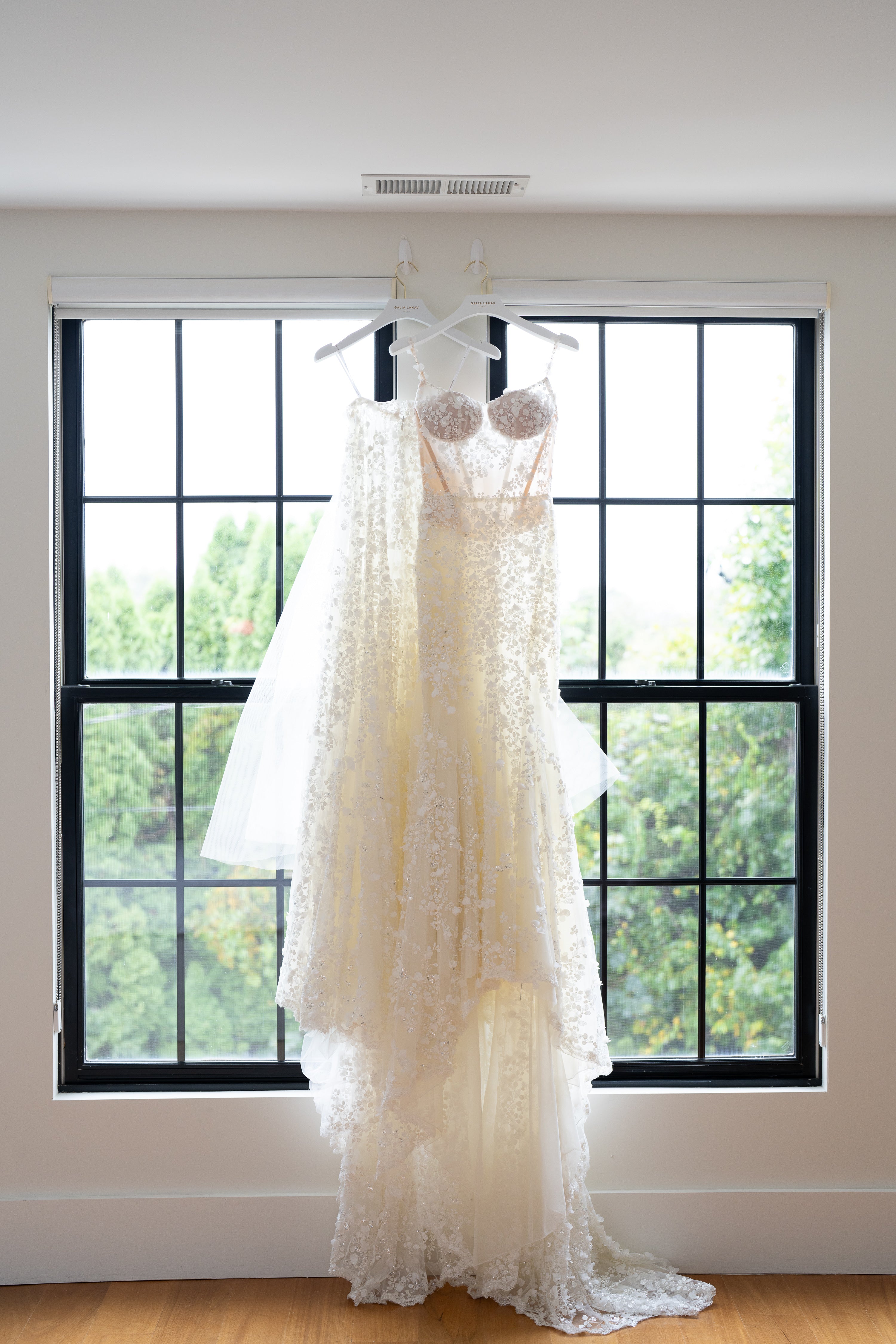 White lace wedding dress hanging in front of a window with a view of trees.