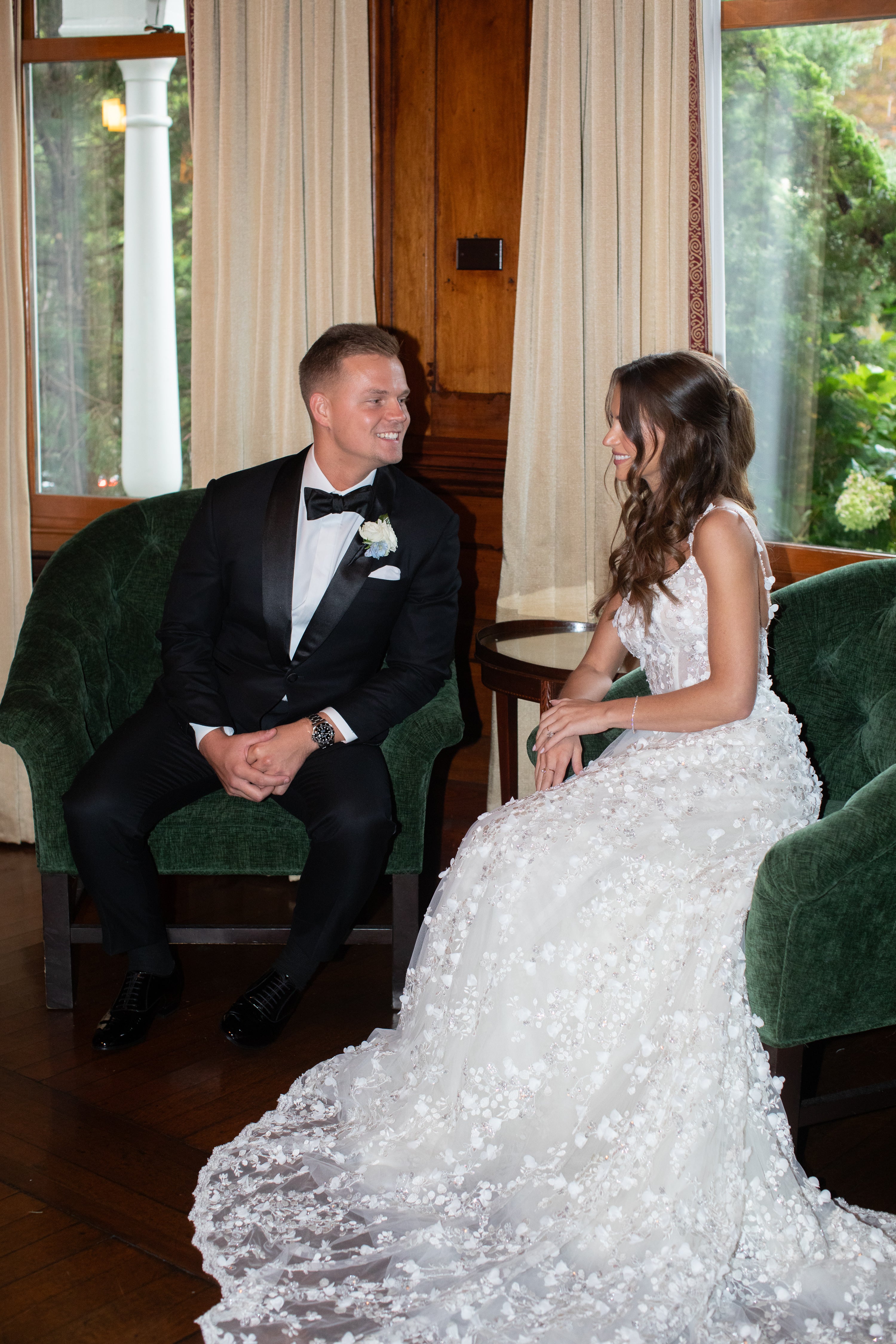 Man in a tuxedo and woman in a white dress sitting on green chairs in a room with large windows.
