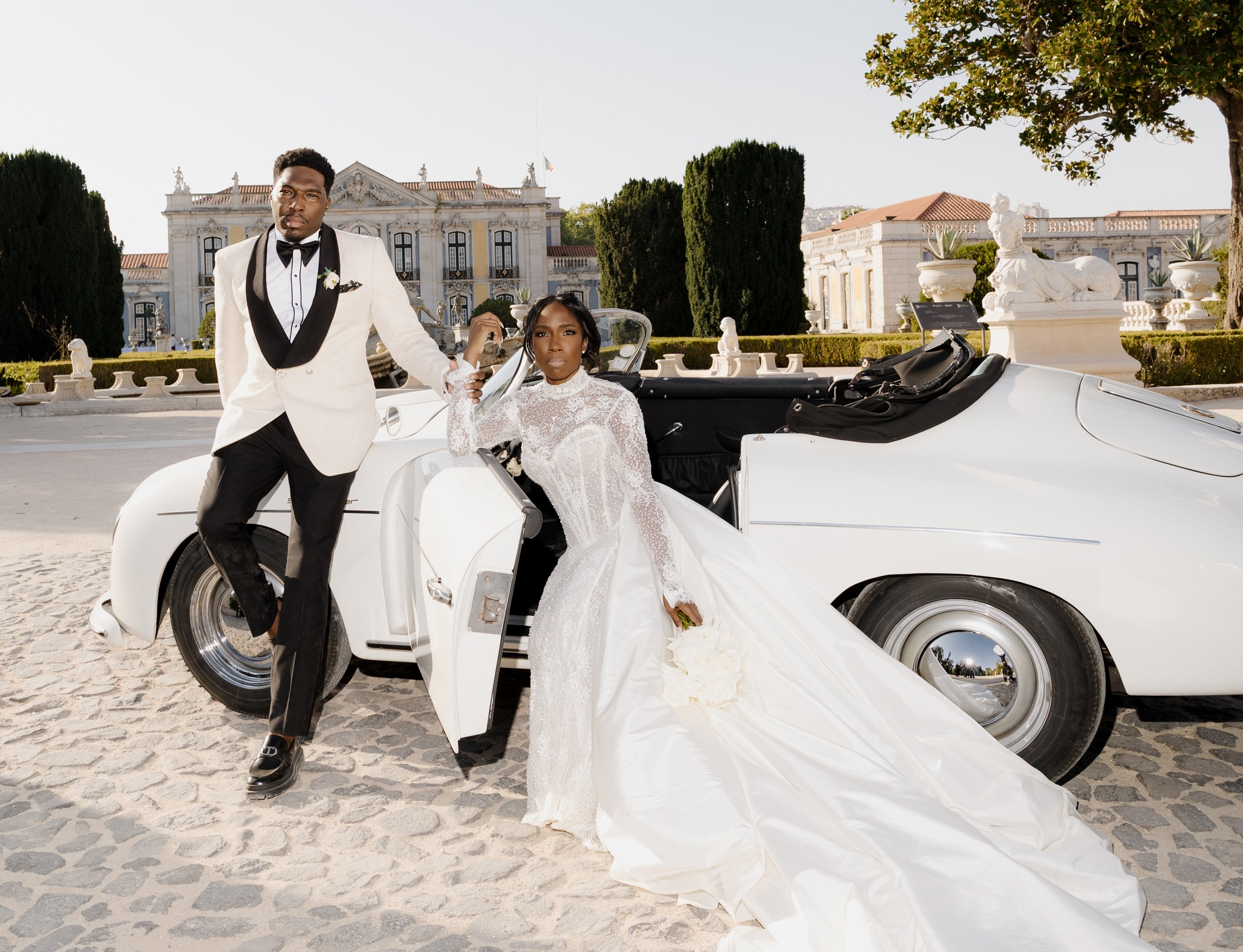 Man and woman in formal attire standing next to a vintage car in an elegant outdoor setting.