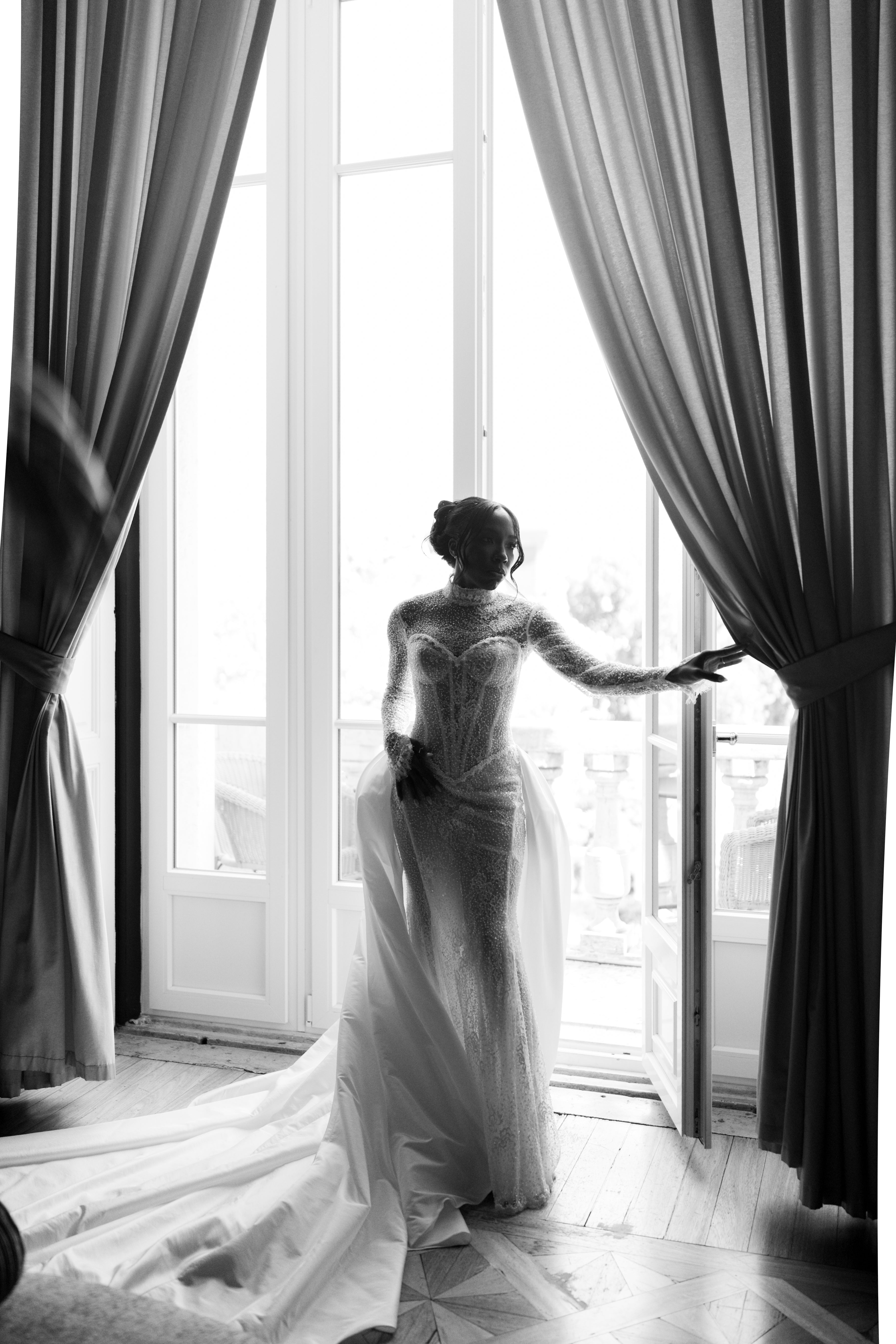 Black and white photo of a woman in a wedding dress standing by a large window with curtains.
