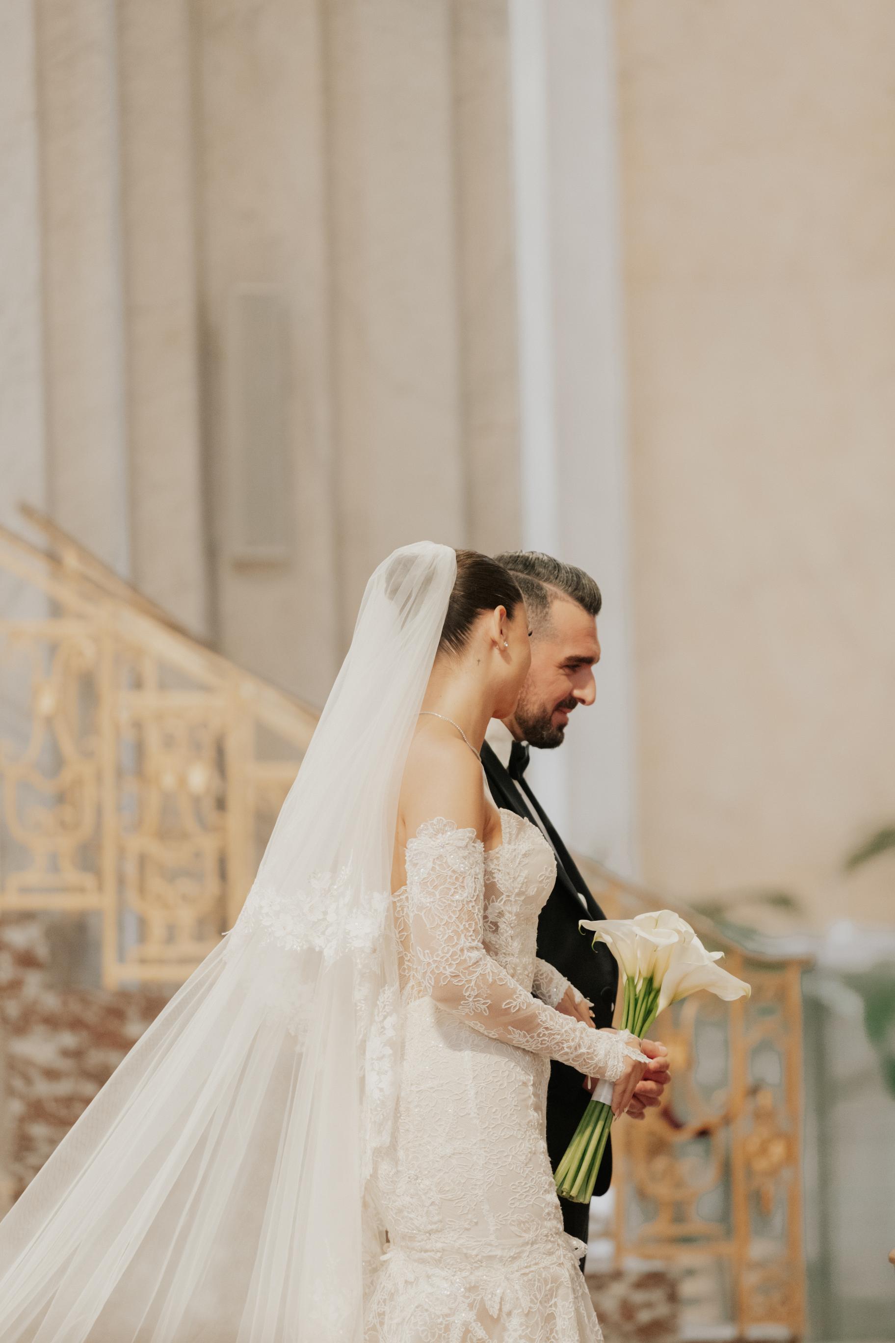 Man and woman in wedding attire with a veil, holding flowers indoors.