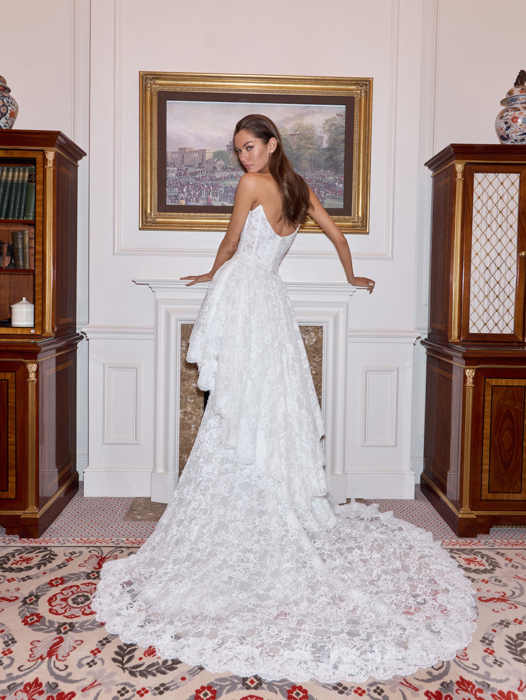 Woman in a white wedding dress standing in an elegant room with wooden furniture and a painting on the wall.
