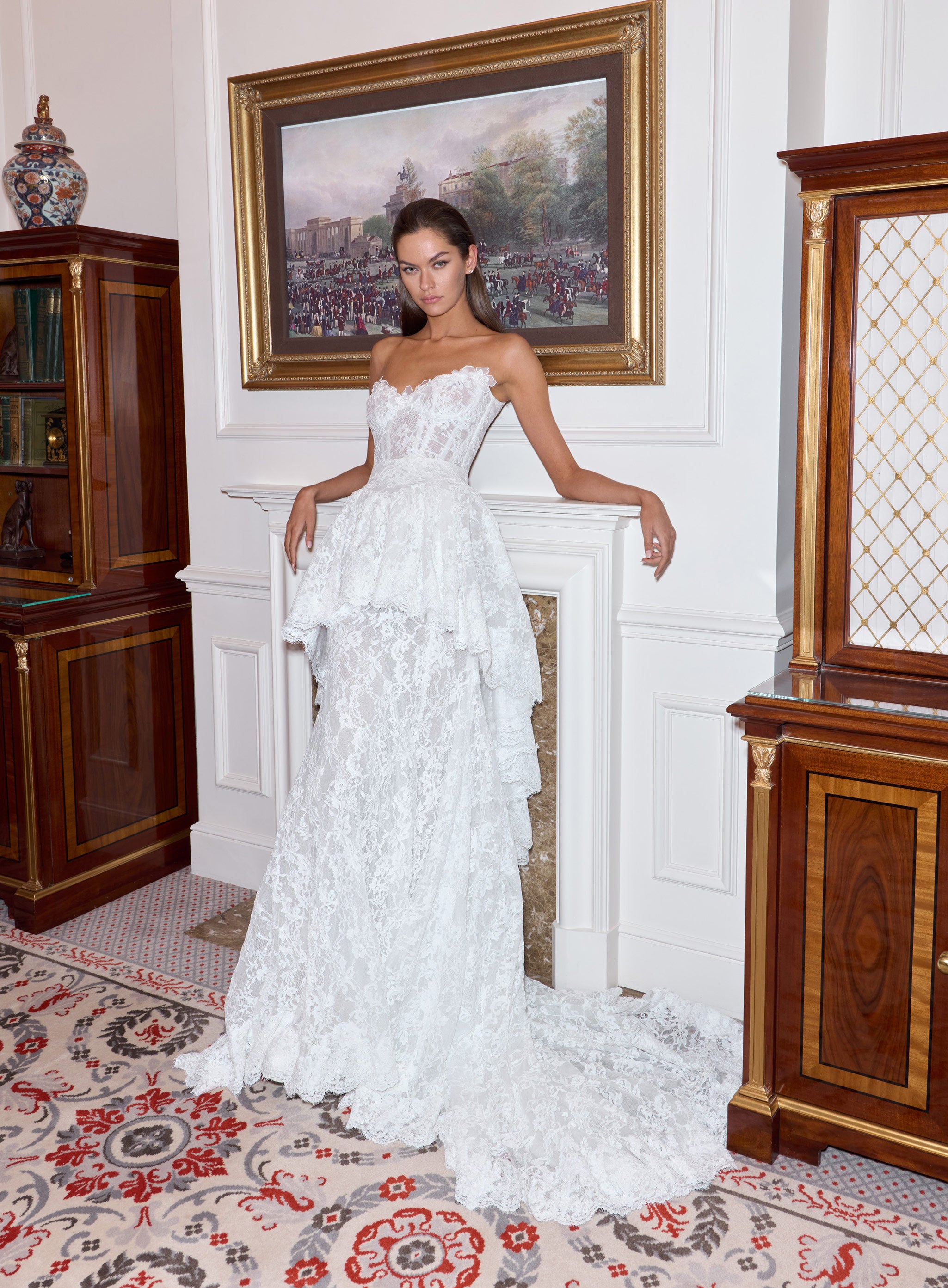 Woman in a white lace dress standing in an elegant room with wooden furniture and a painting on the wall.