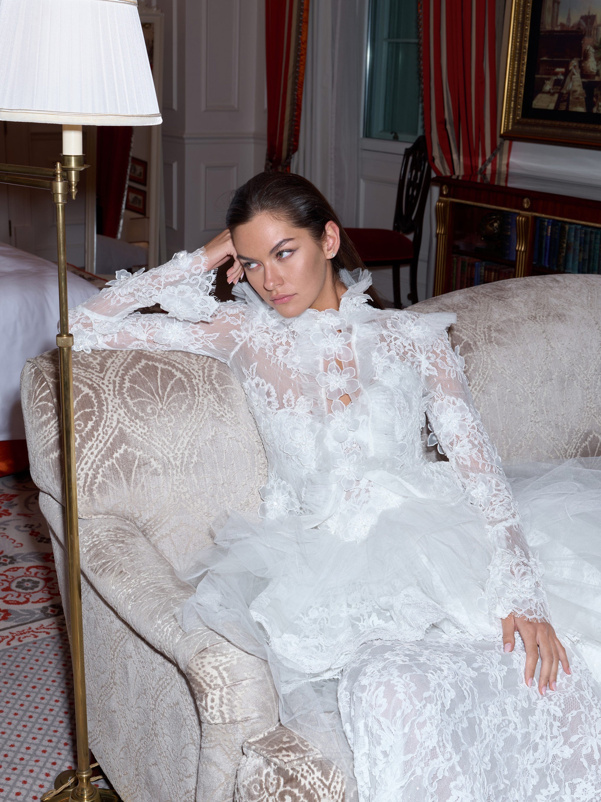 Woman in a white lace dress sitting on a patterned sofa in an elegant room.