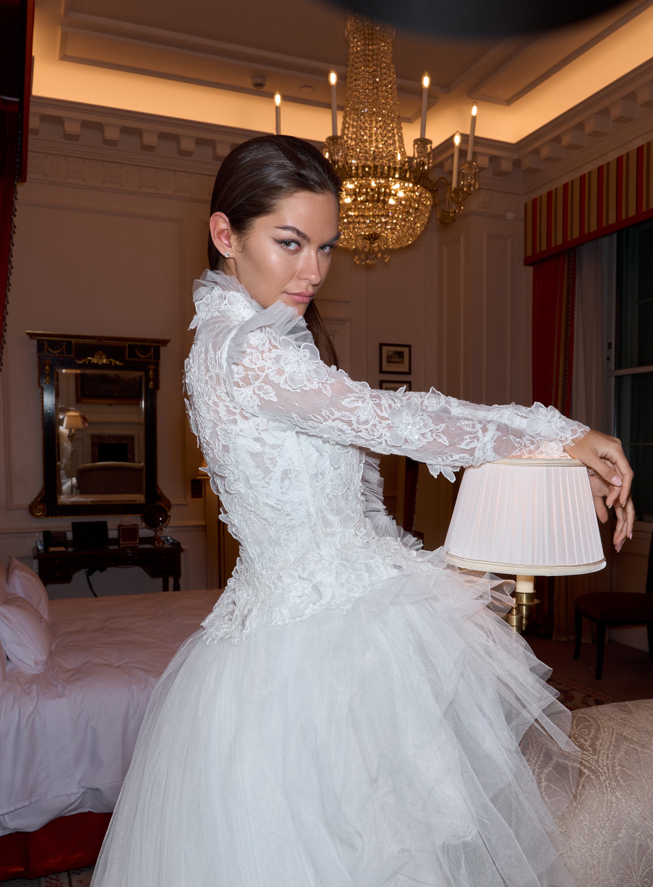 Woman in a white lace dress standing in a luxurious room with a chandelier.
