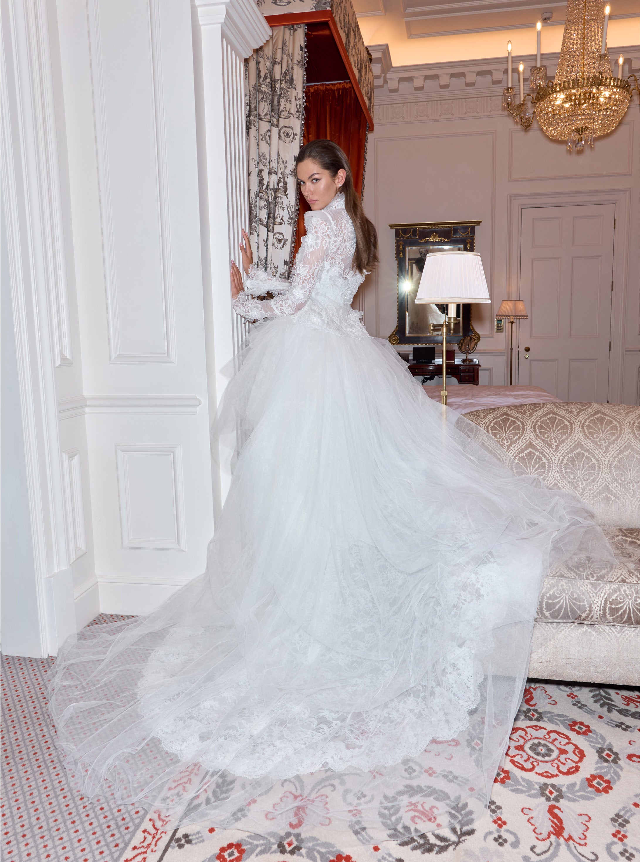 Woman in a white wedding dress standing in an elegant room with chandelier and decor.