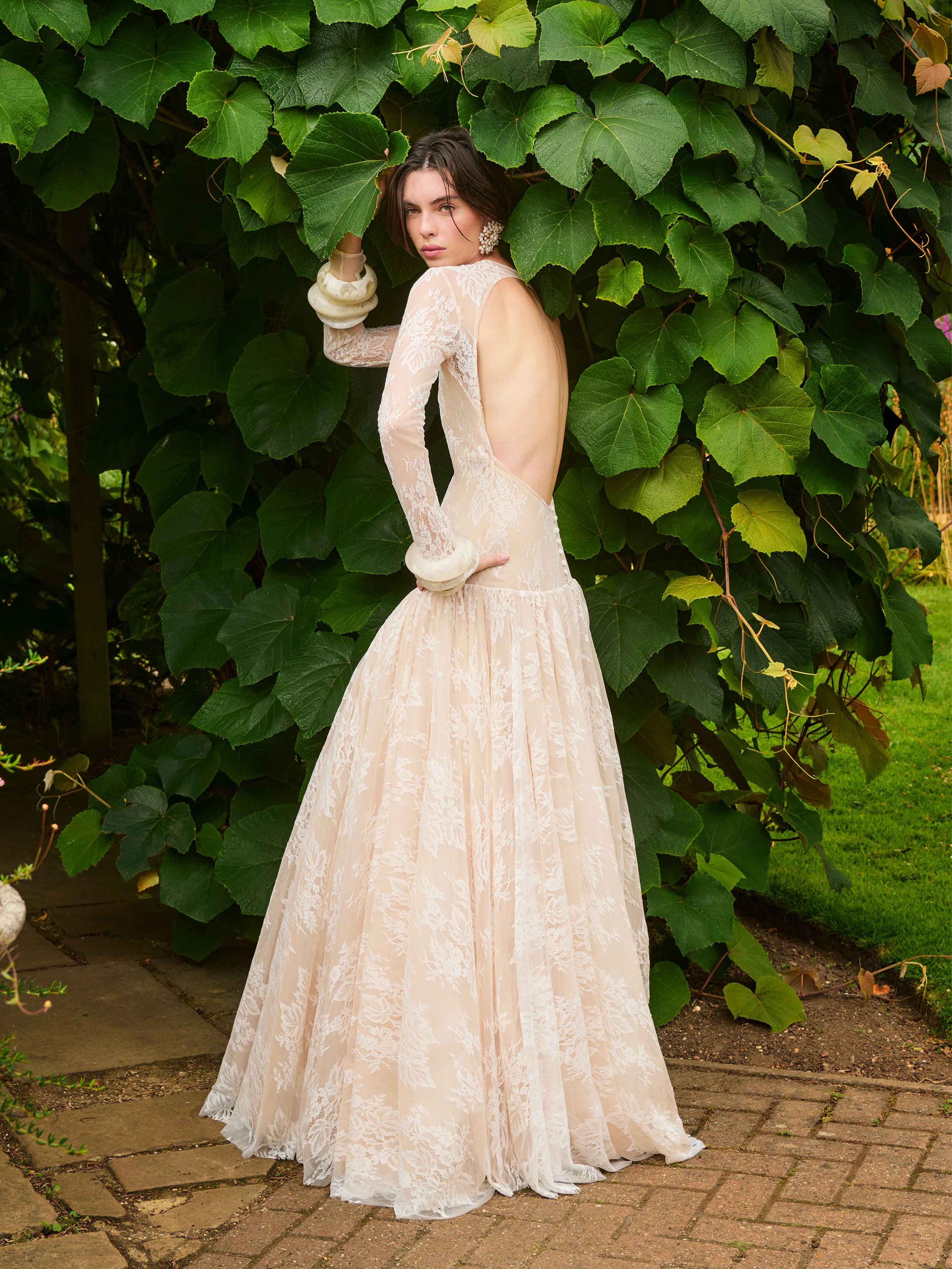 Woman in a beige lace dress standing in front of a green leafy wall.