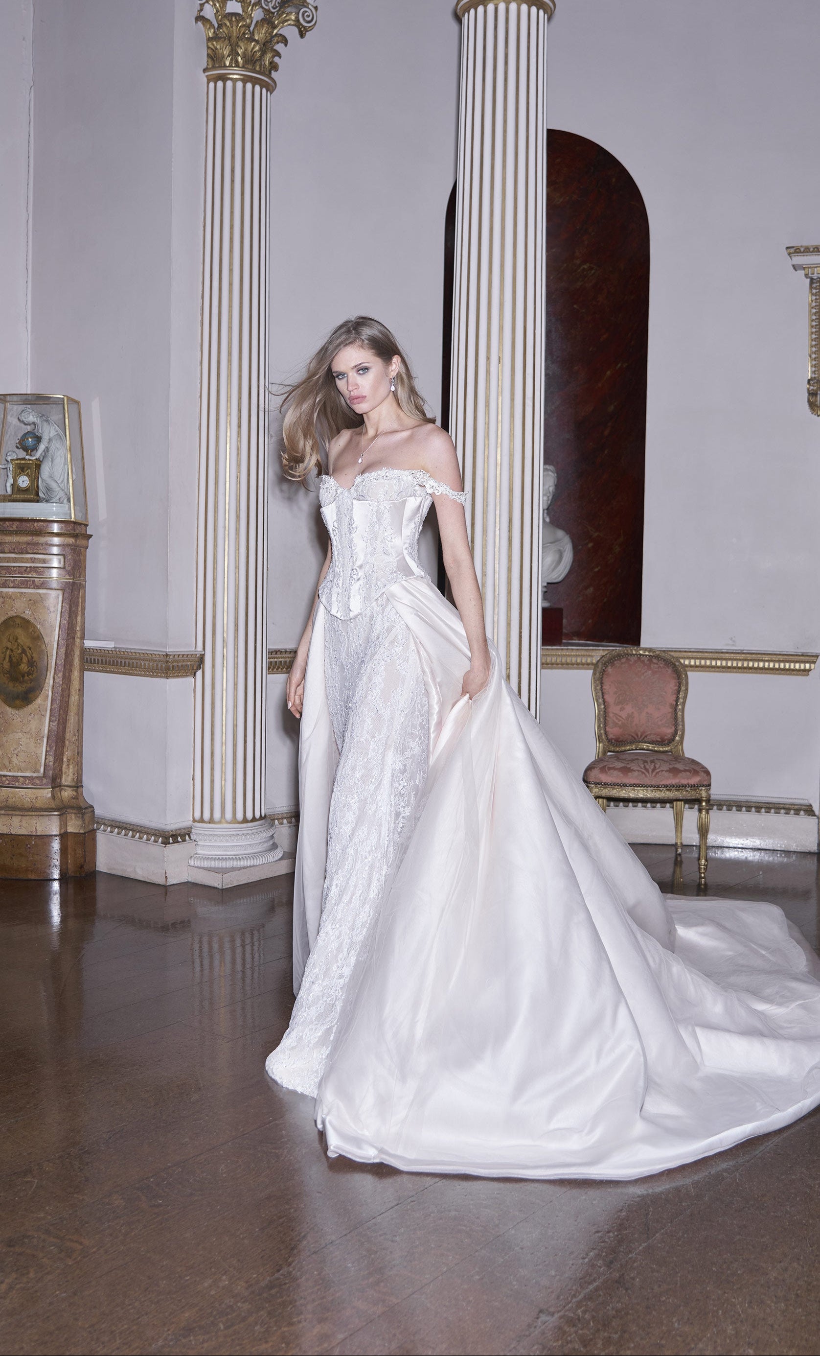 Woman in a white wedding dress standing in an elegant room with columns and a chair.