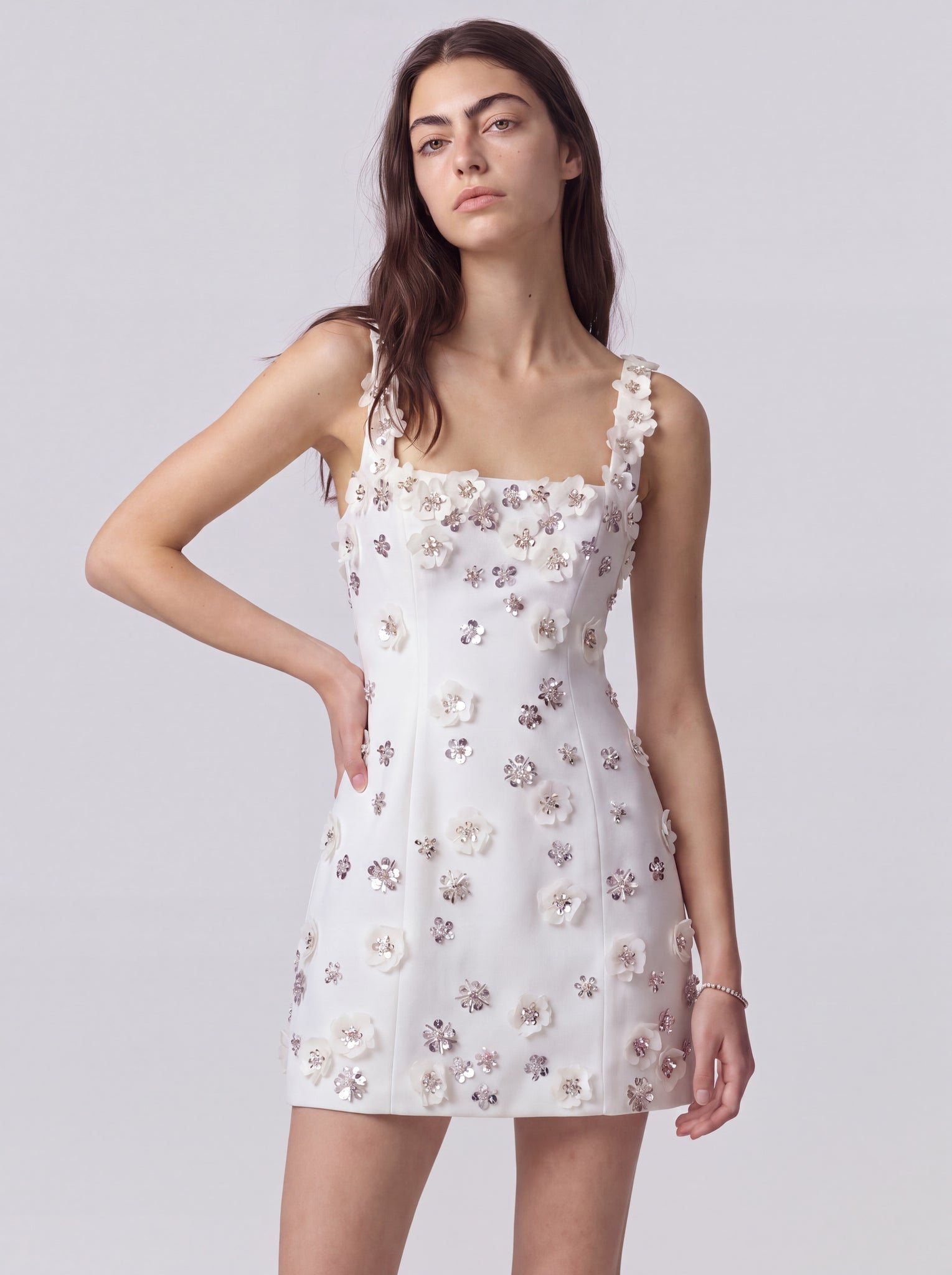 A woman modeling a white sleeveless mini dress with 3D floral appliqués and ruffled shoulder straps, photographed against a solid light gray studio background.