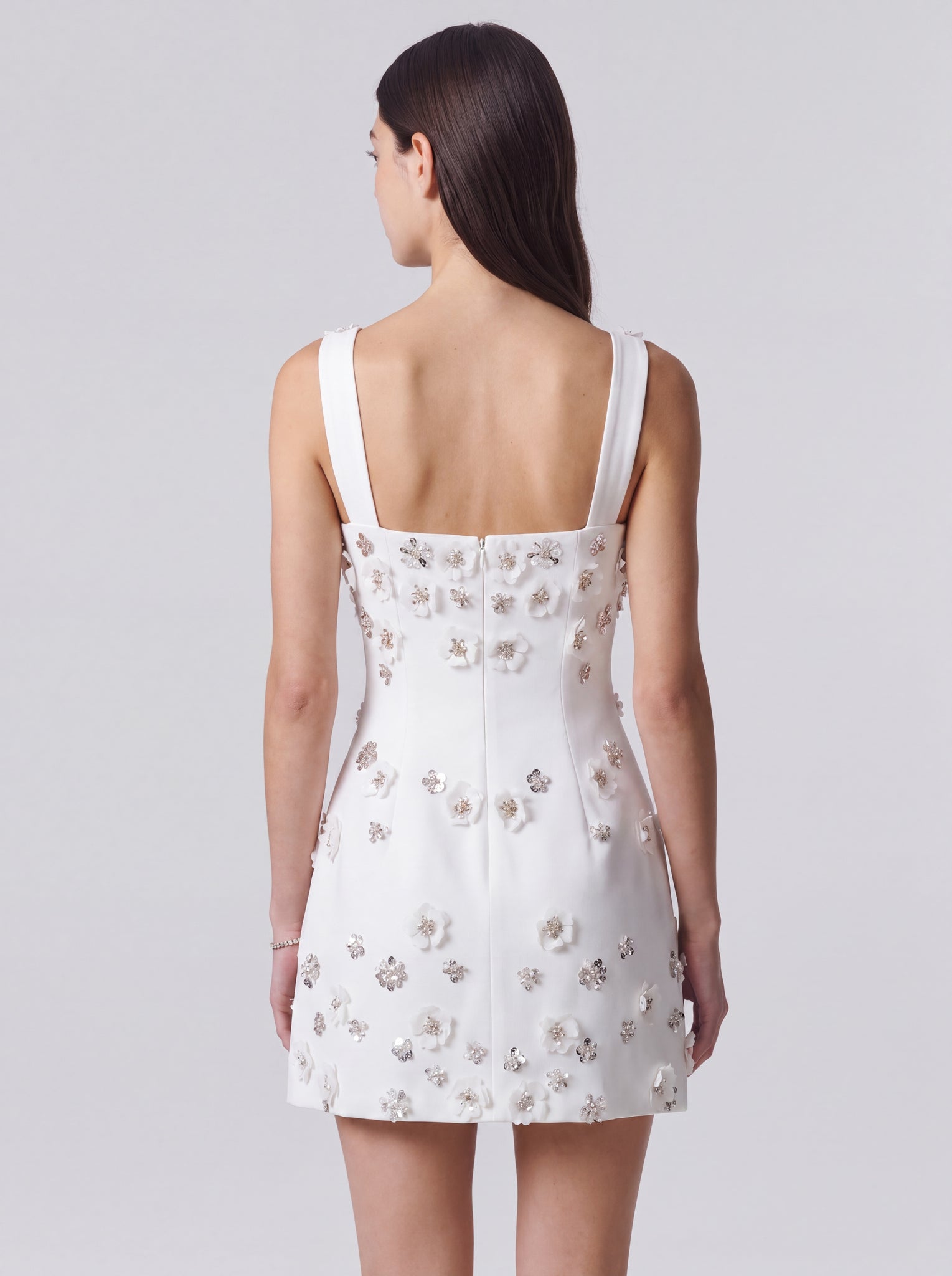 A woman shown from the back wearing a white sleeveless mini dress with floral applique and a square neckline, photographed against a light gray studio background.