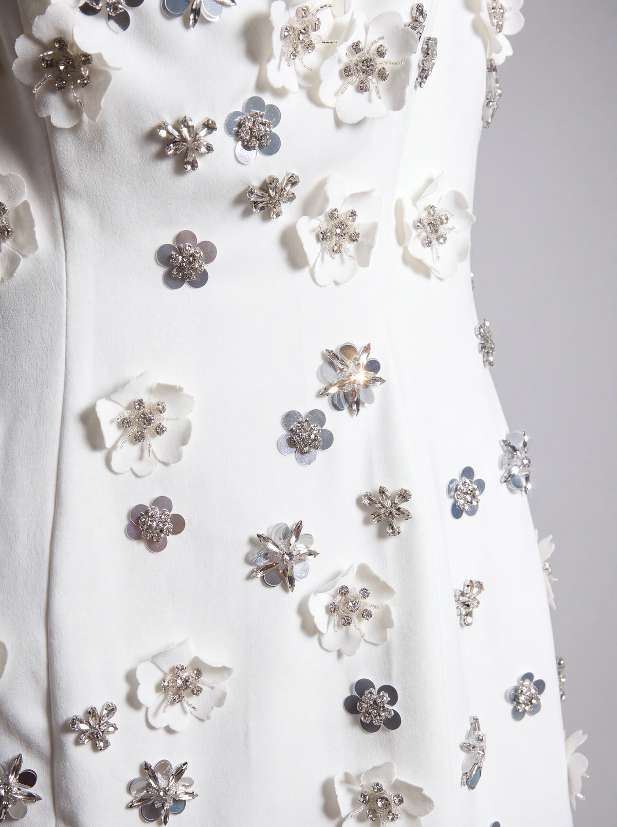 Close-up of a white garment featuring 3D floral appliqués in white and silver with rhinestone centers, displayed on a mannequin or dress form against a light gray background.