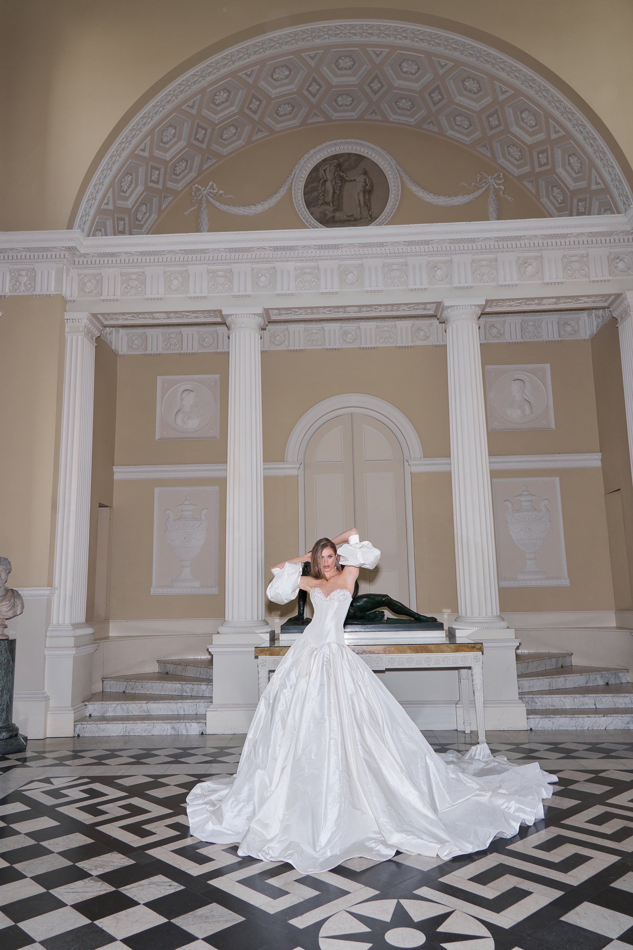 Woman in a white wedding dress standing in a grand hall with decorative columns and ceiling.