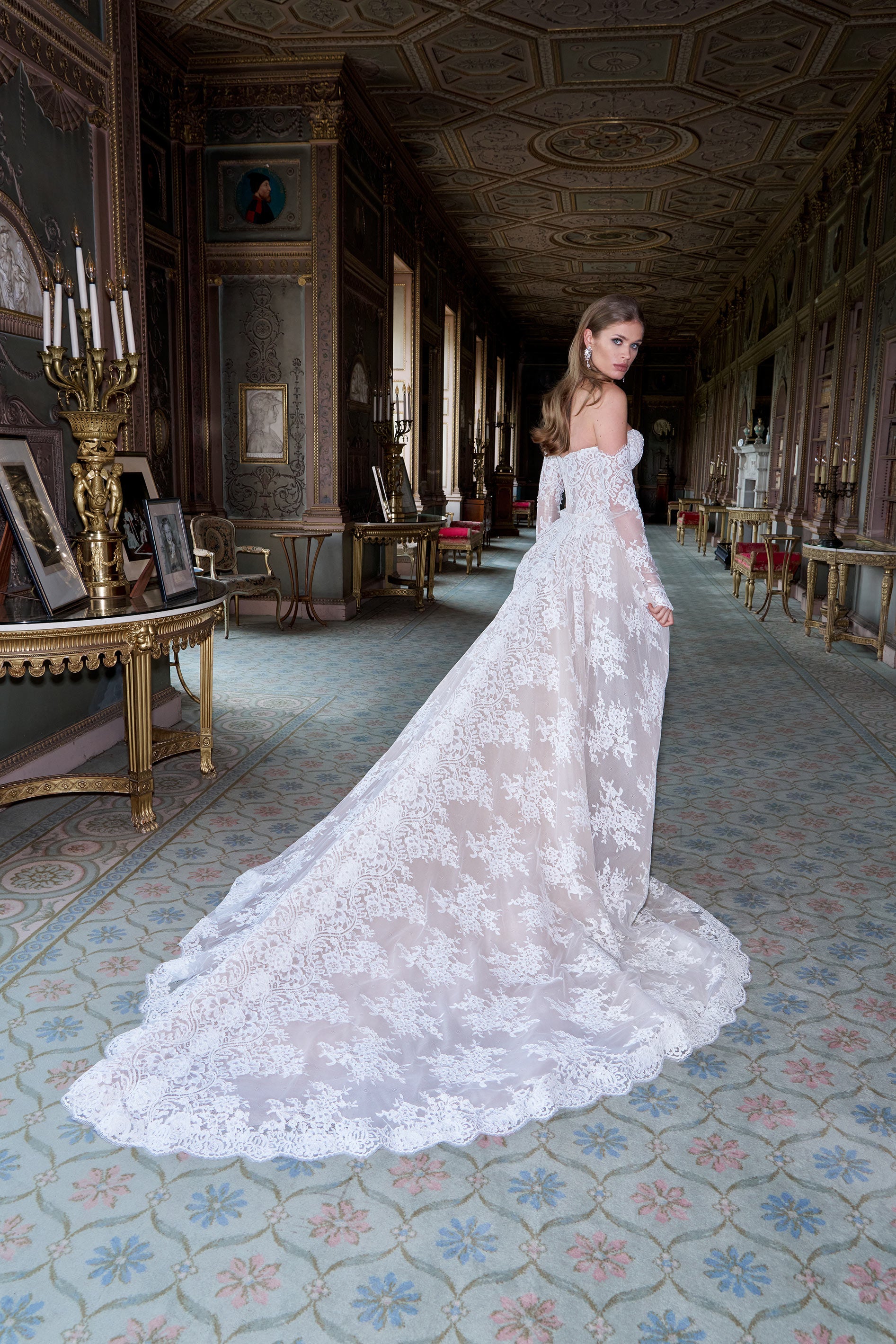 Woman in a white lace wedding dress standing in an elegant hallway with decorative ceiling and floor.