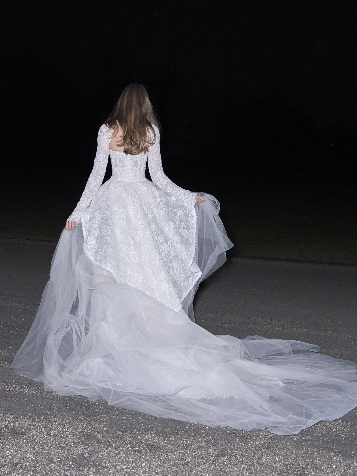 Woman in a white wedding dress with a long train standing on a dark road.