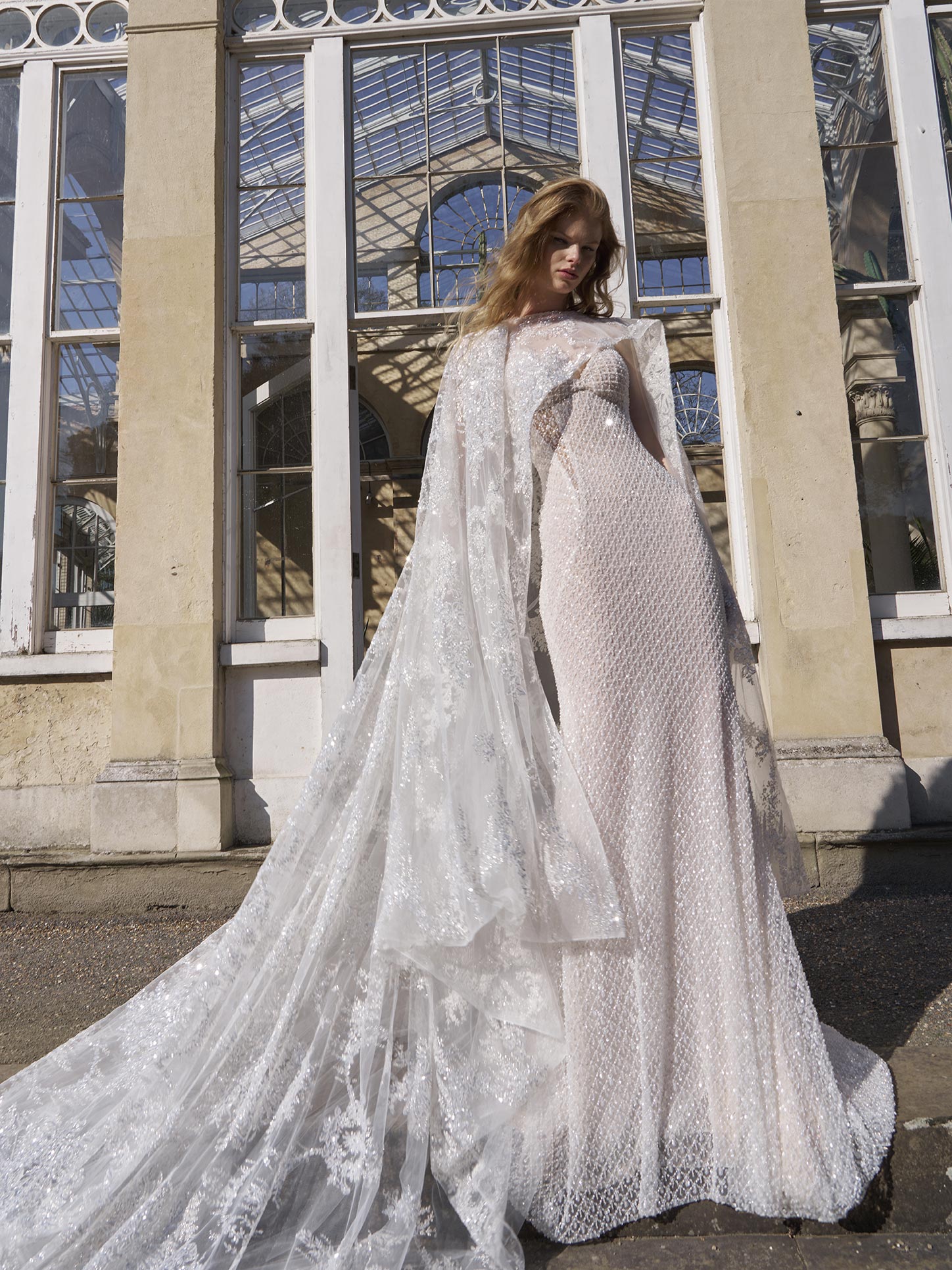 Woman wearing a white lace wedding dress with a long train in front of a glass building.