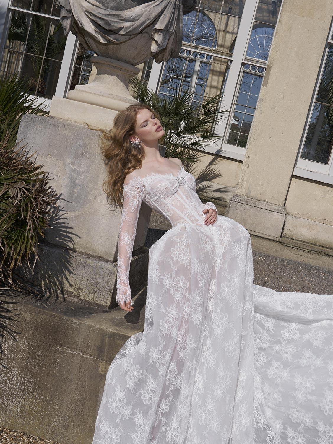 Woman in a white lace wedding dress standing outdoors near classical architecture.