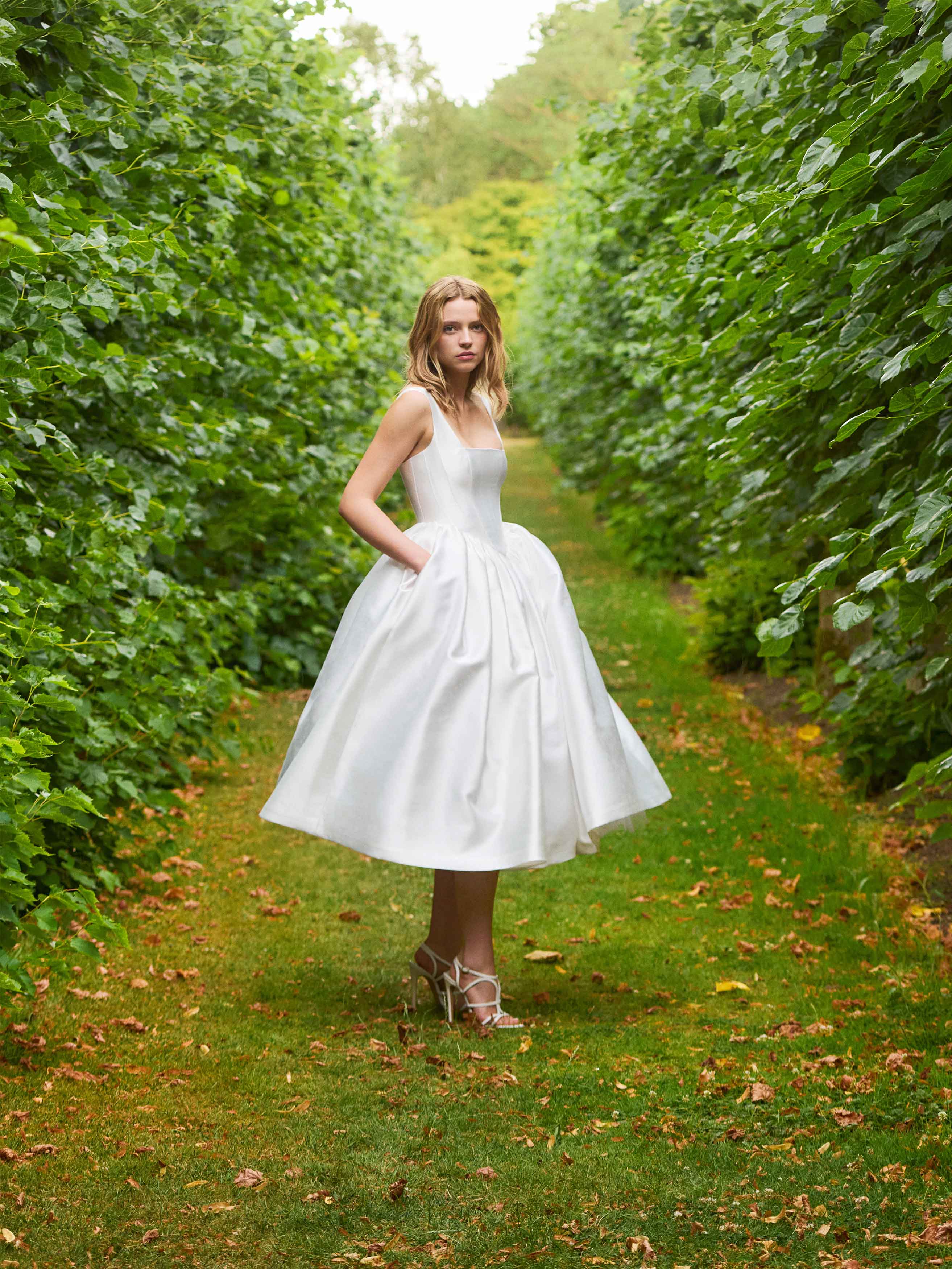 Woman in a white dress standing between two rows of green hedges