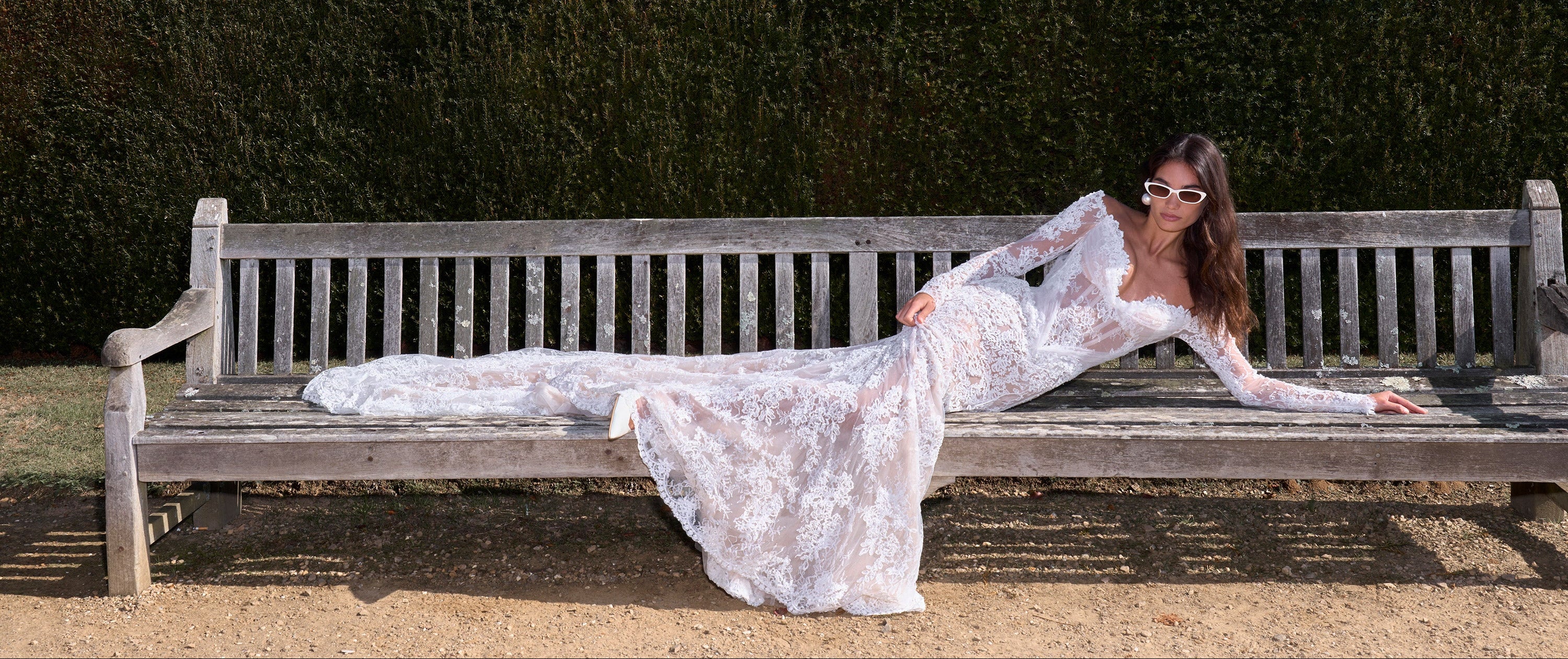 Woman in a white lace dress lying on a wooden bench against a hedge.