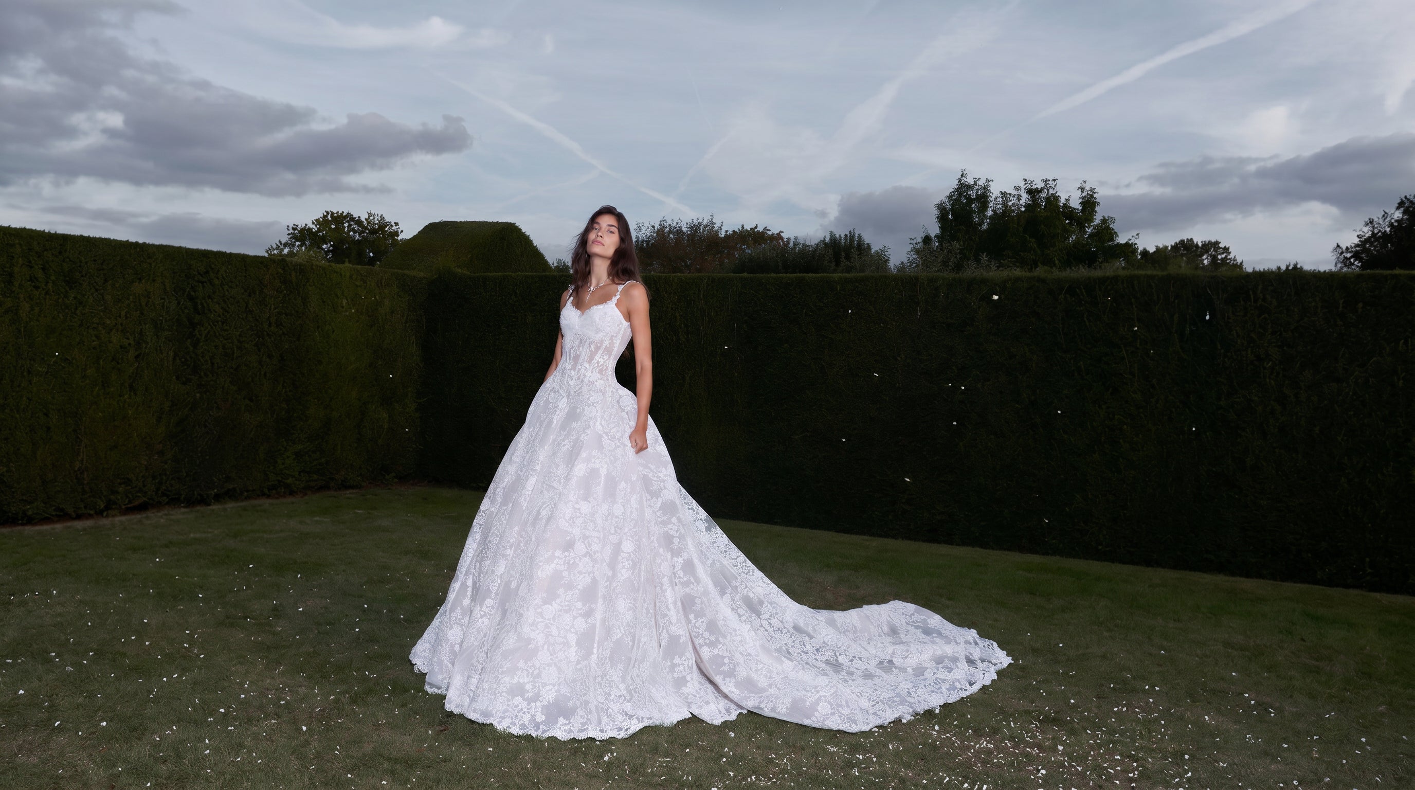 Woman in a white wedding dress standing in a garden with dark hedges and a cloudy sky.