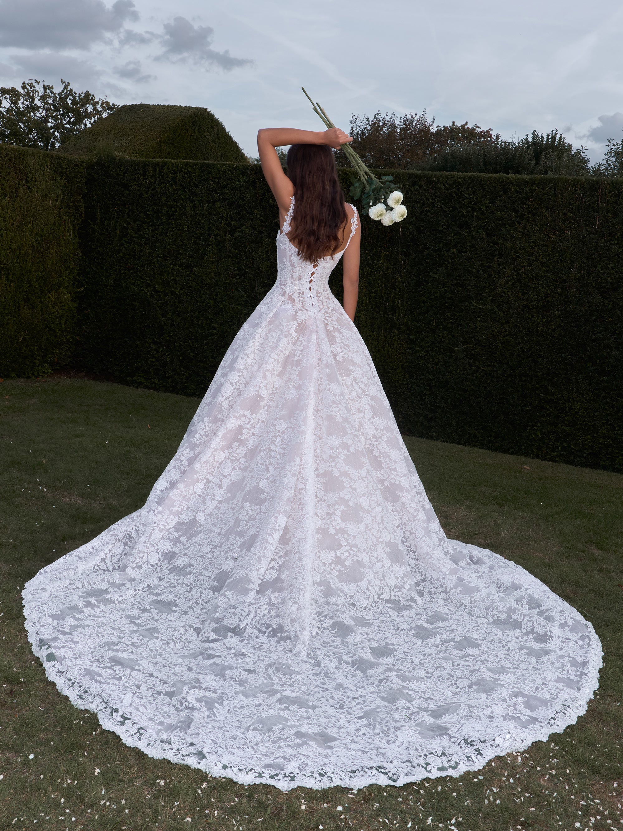 Woman in a white lace wedding dress holding flowers in an outdoor setting.