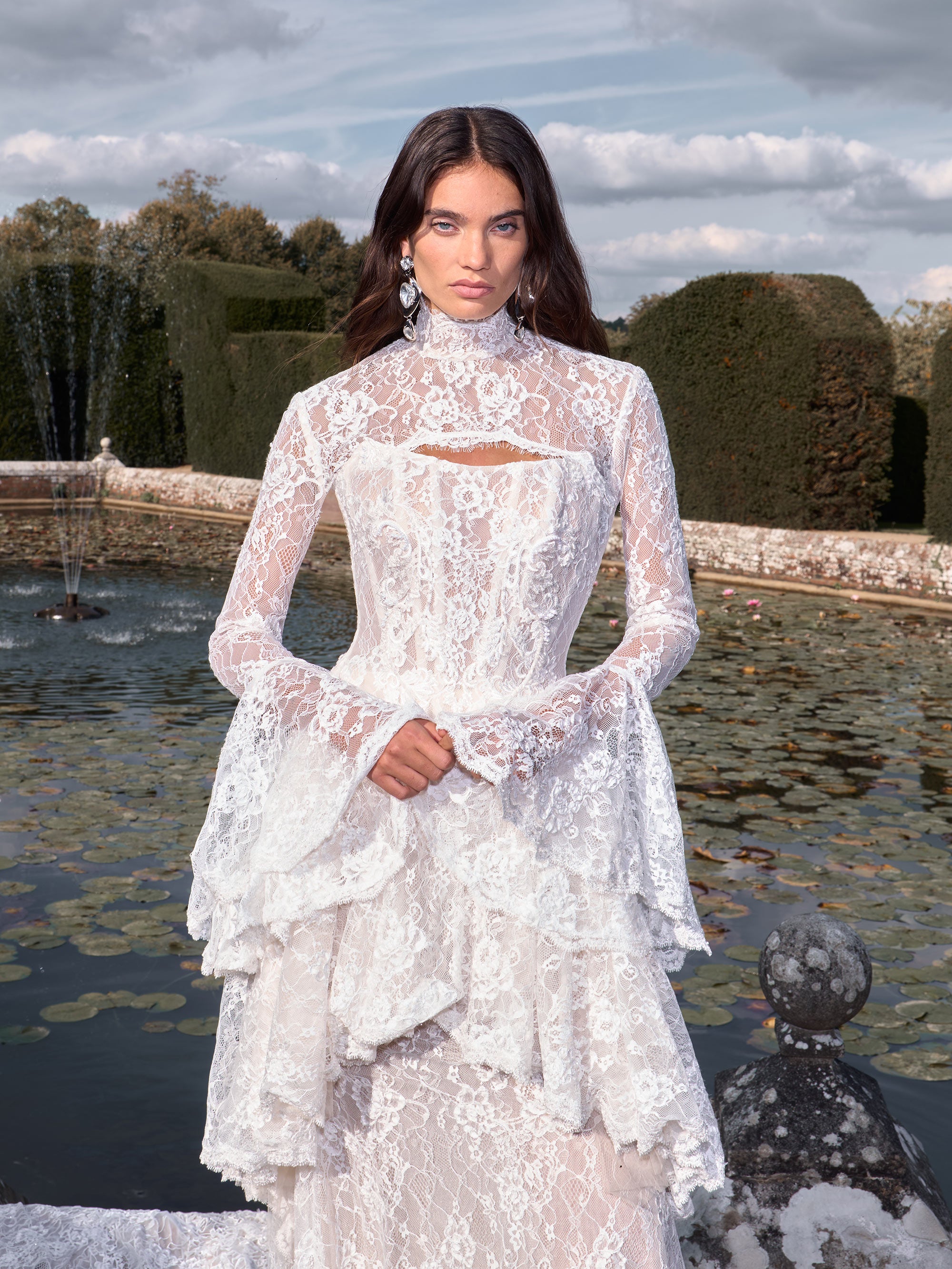 Woman in a white lace dress standing in front of a fountain with trees and clouds in the background