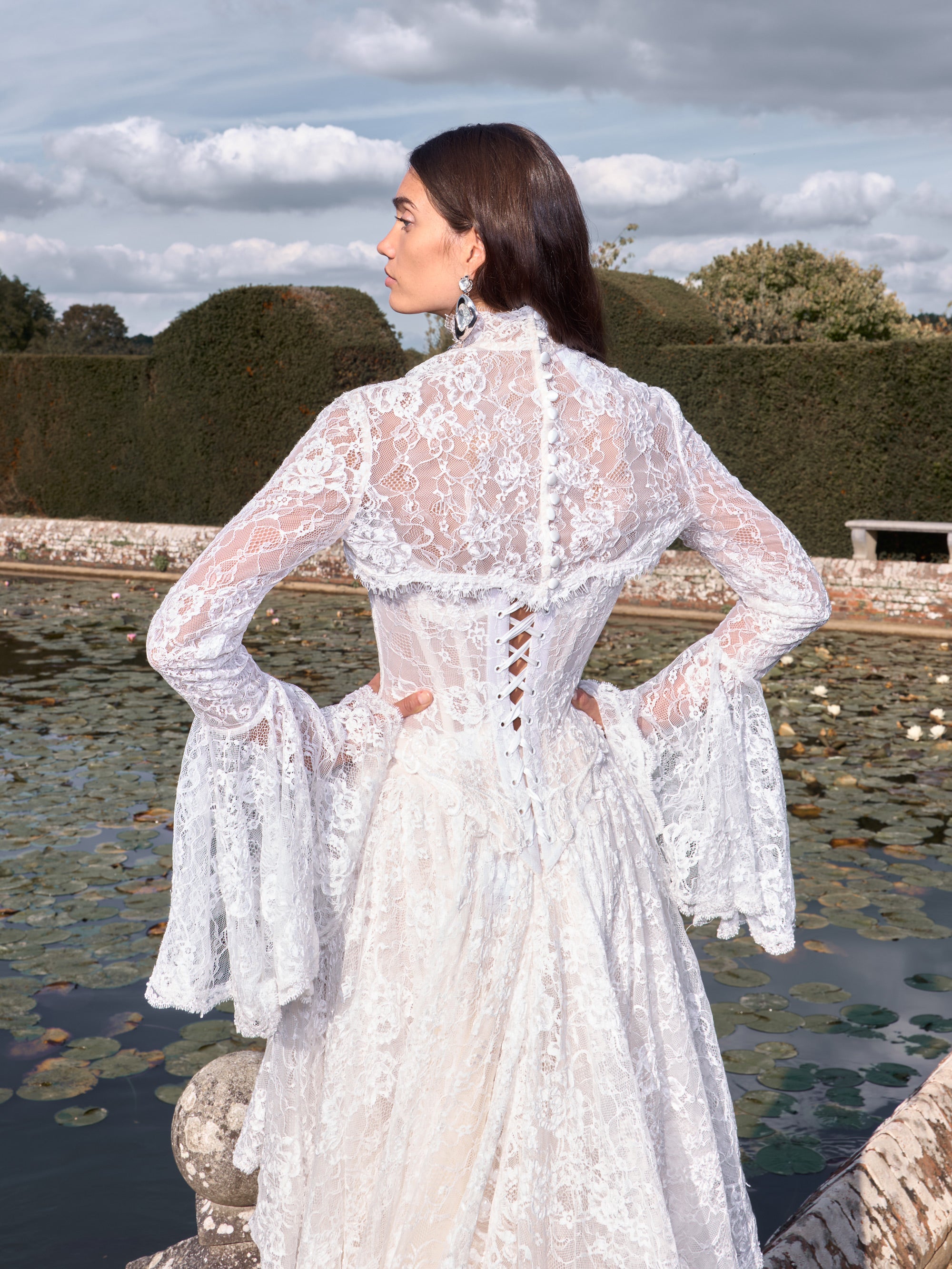 Woman in a white lace dress standing by a pond with greenery in the background