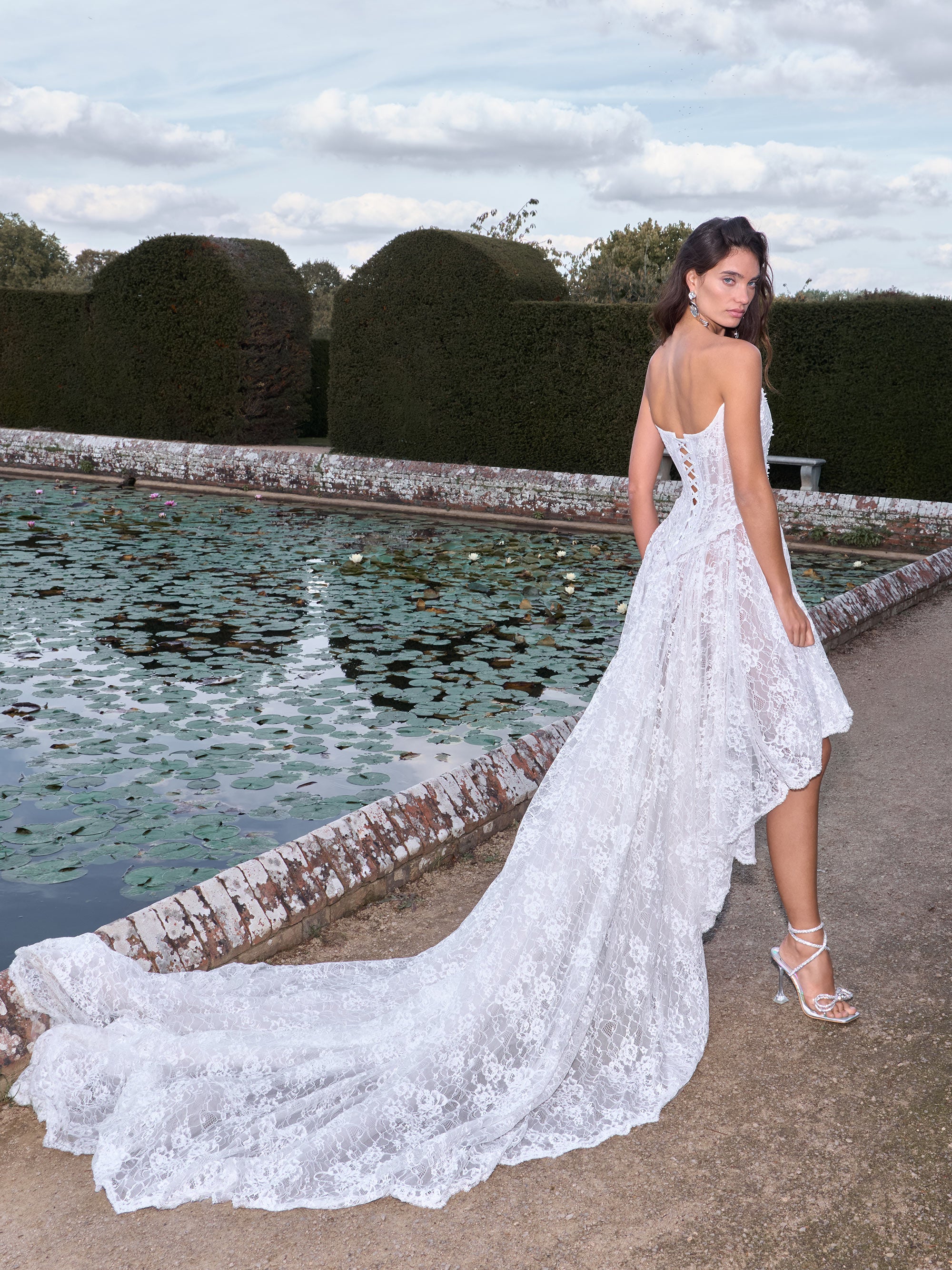Woman in a white lace dress standing by a pond with greenery in the background