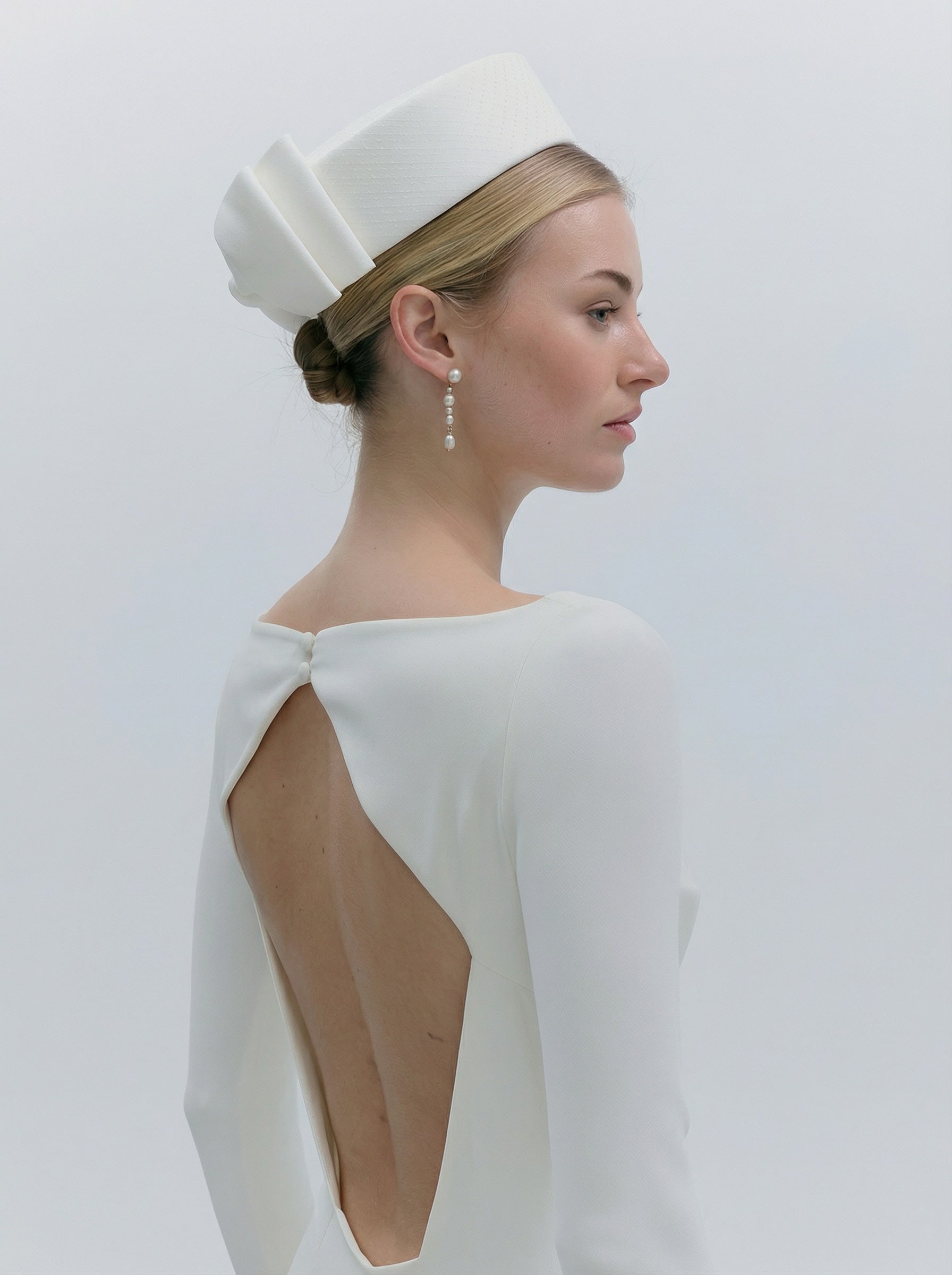 Woman wearing a white dress with a cutout back and a matching headpiece against a plain background