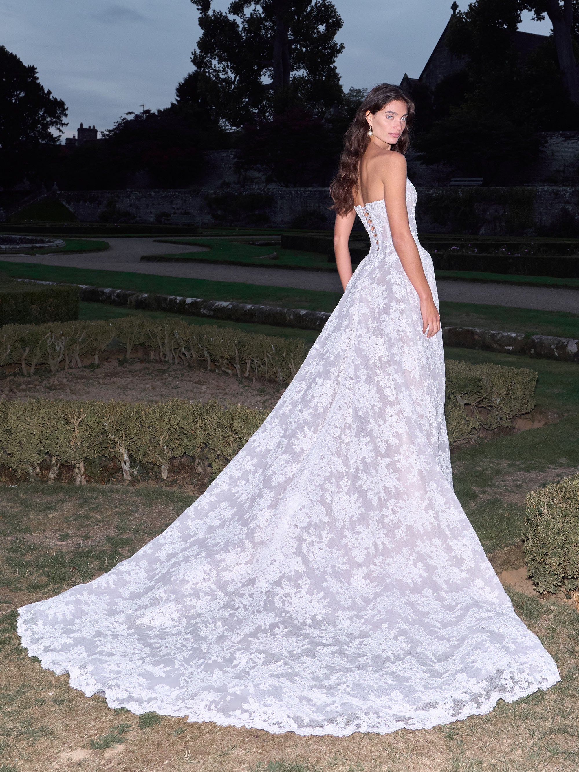 Woman in a white lace wedding dress standing in a garden at dusk.