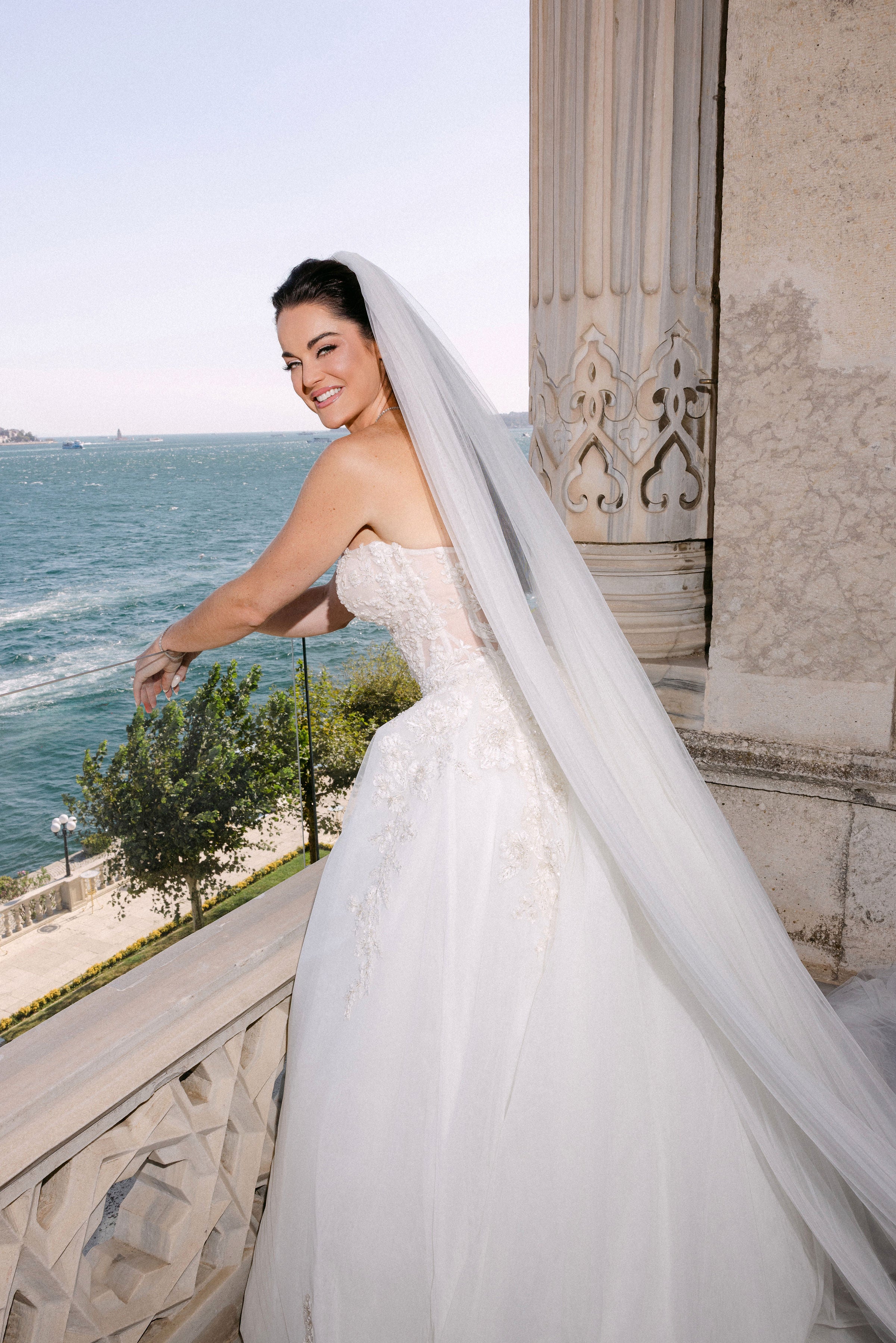 Bride in a white wedding dress with a long veil standing by a waterfront.