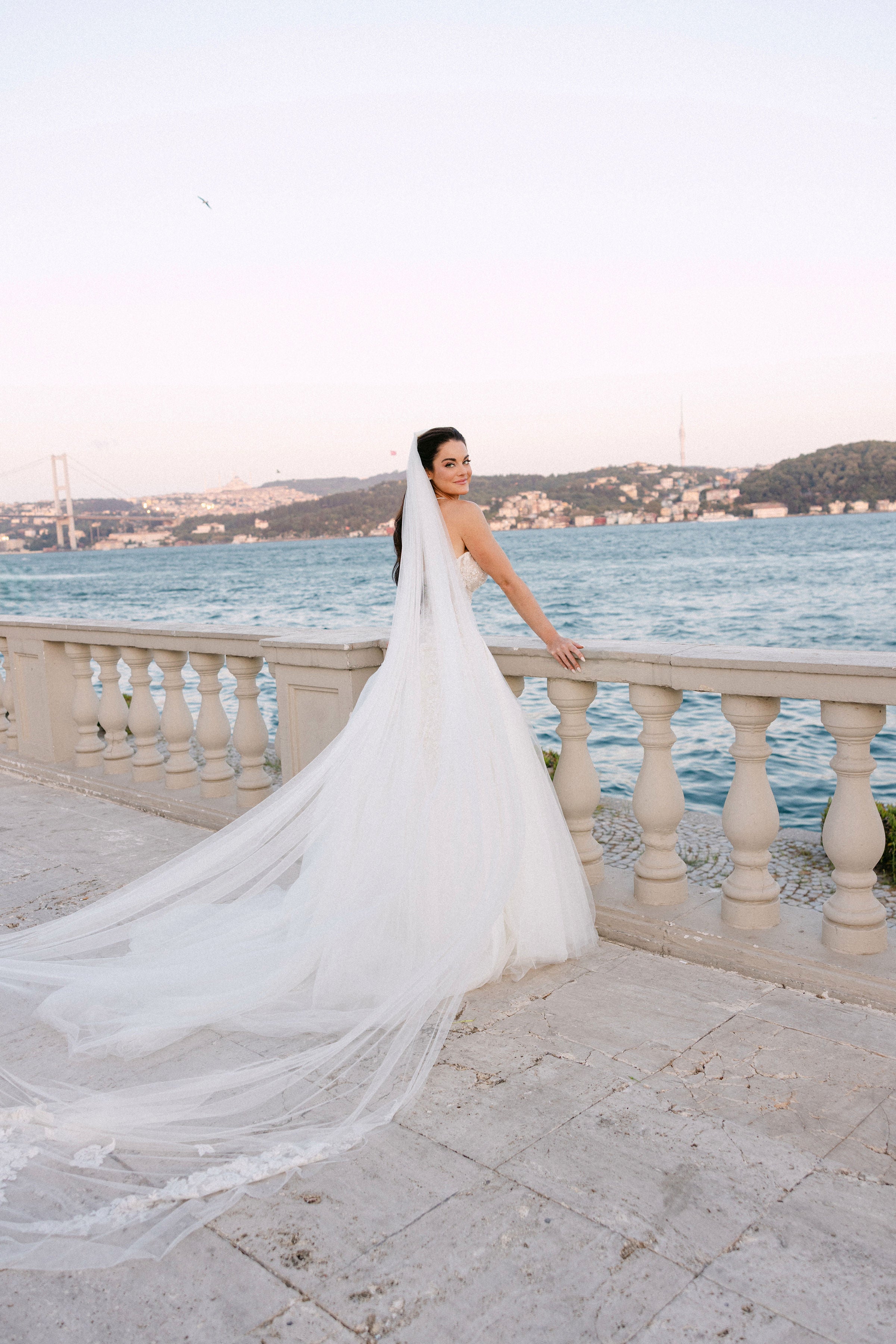 Woman in a white wedding dress with a long veil standing by a waterfront.