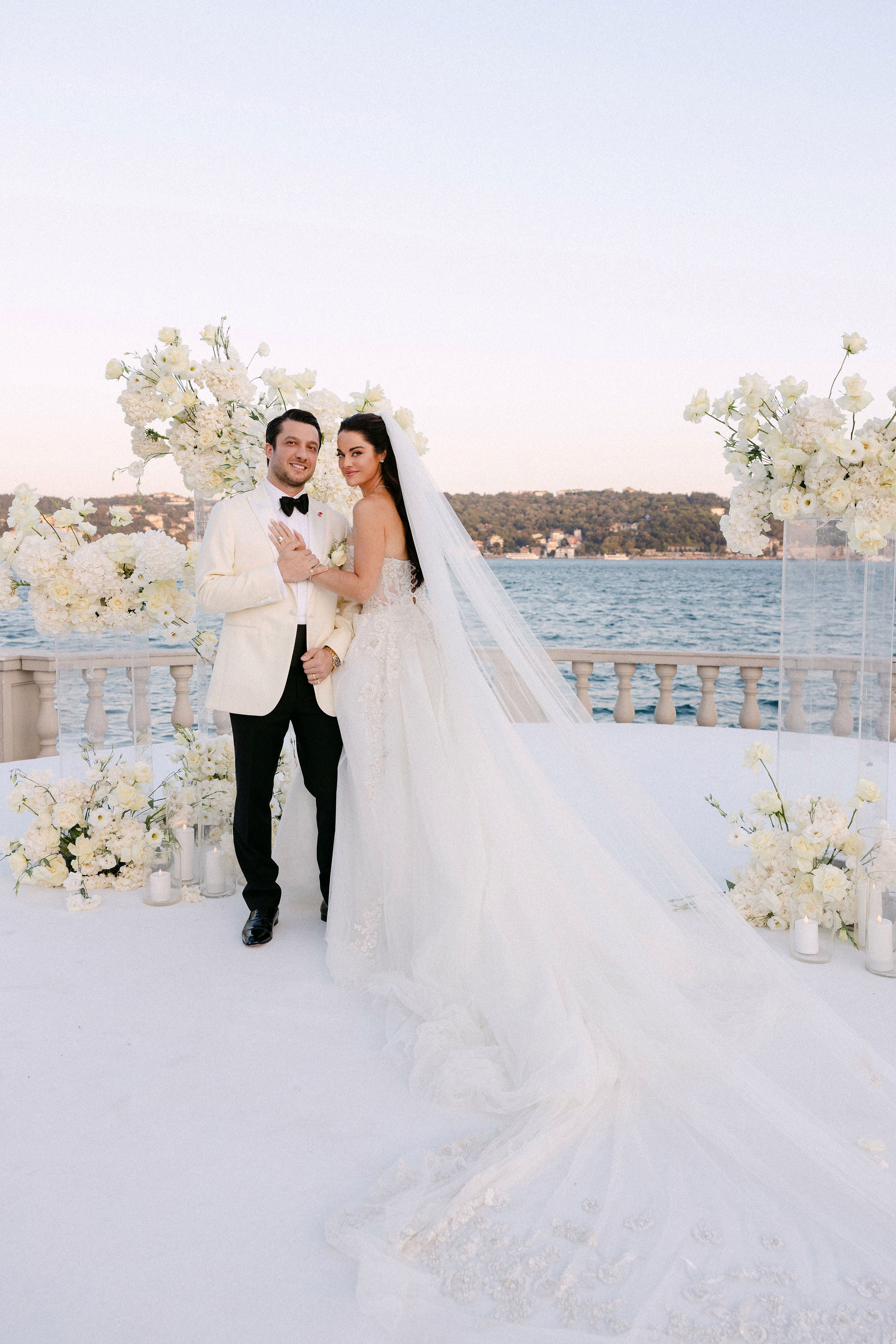 Wedding couple standing in front of a scenic backdrop with flowers and water.