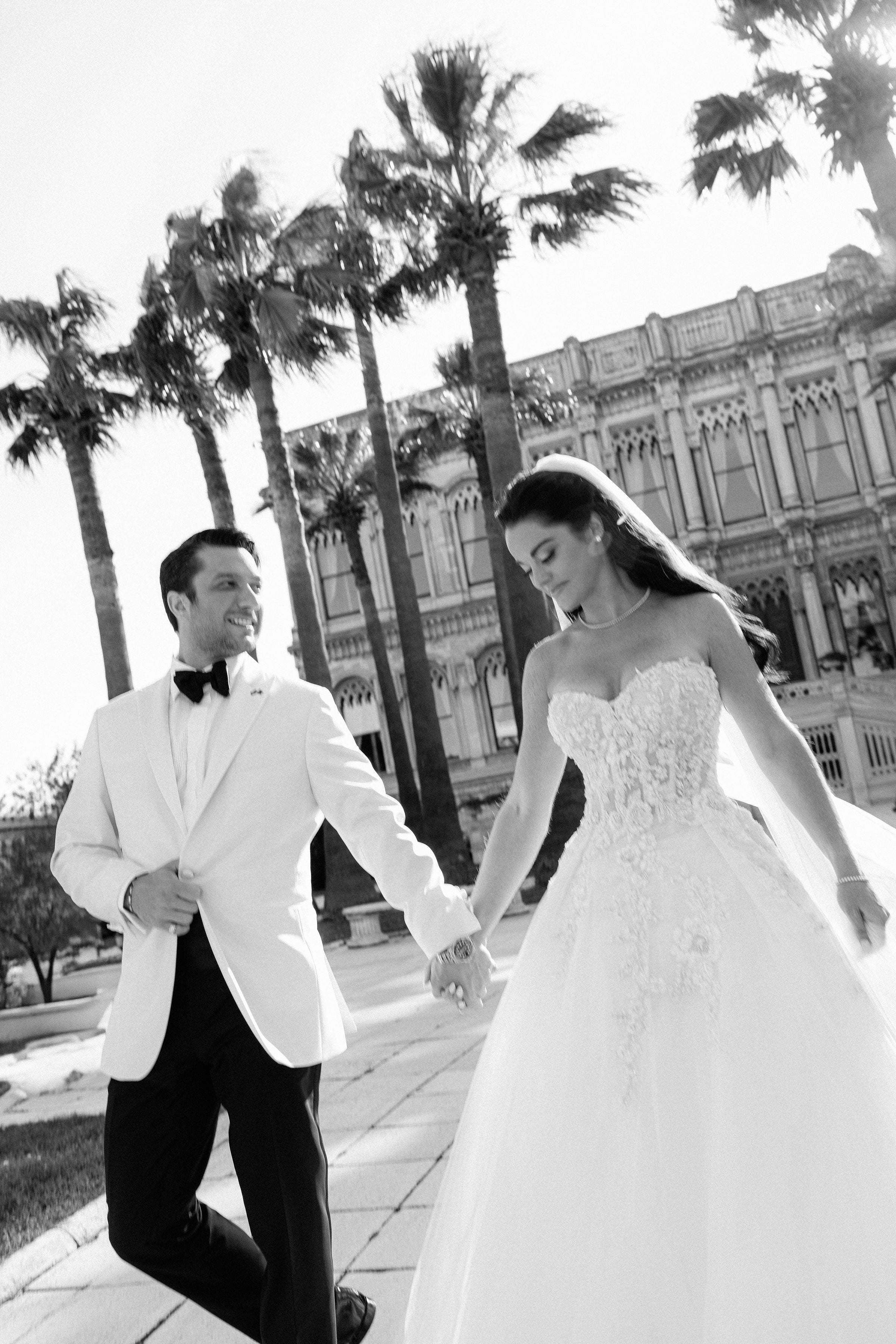 Black and white photo of a bride and groom walking together with palm trees and a building in the background.