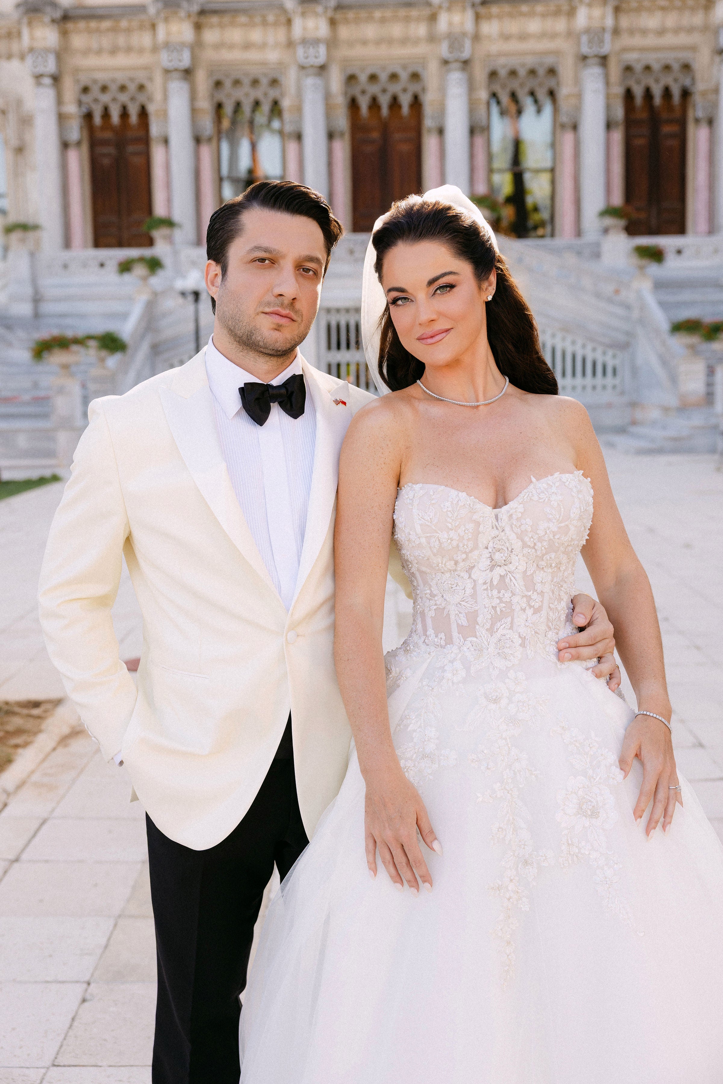 Man in a white tuxedo and woman in a white wedding dress standing together in front of an elegant building.