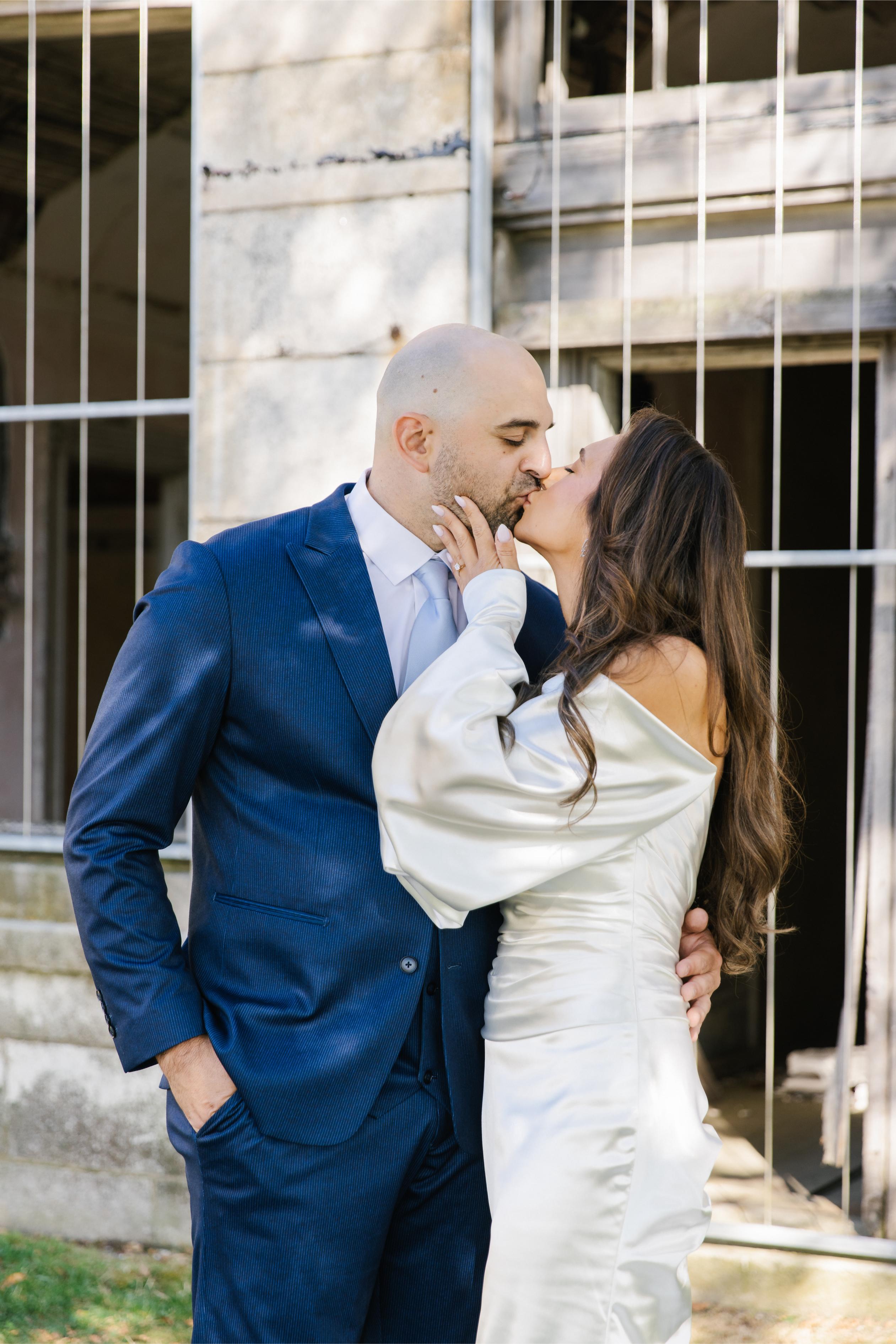 GL Bride Alexandra in Galia Lahav's Alona kissing her husband in front of a stone building.
