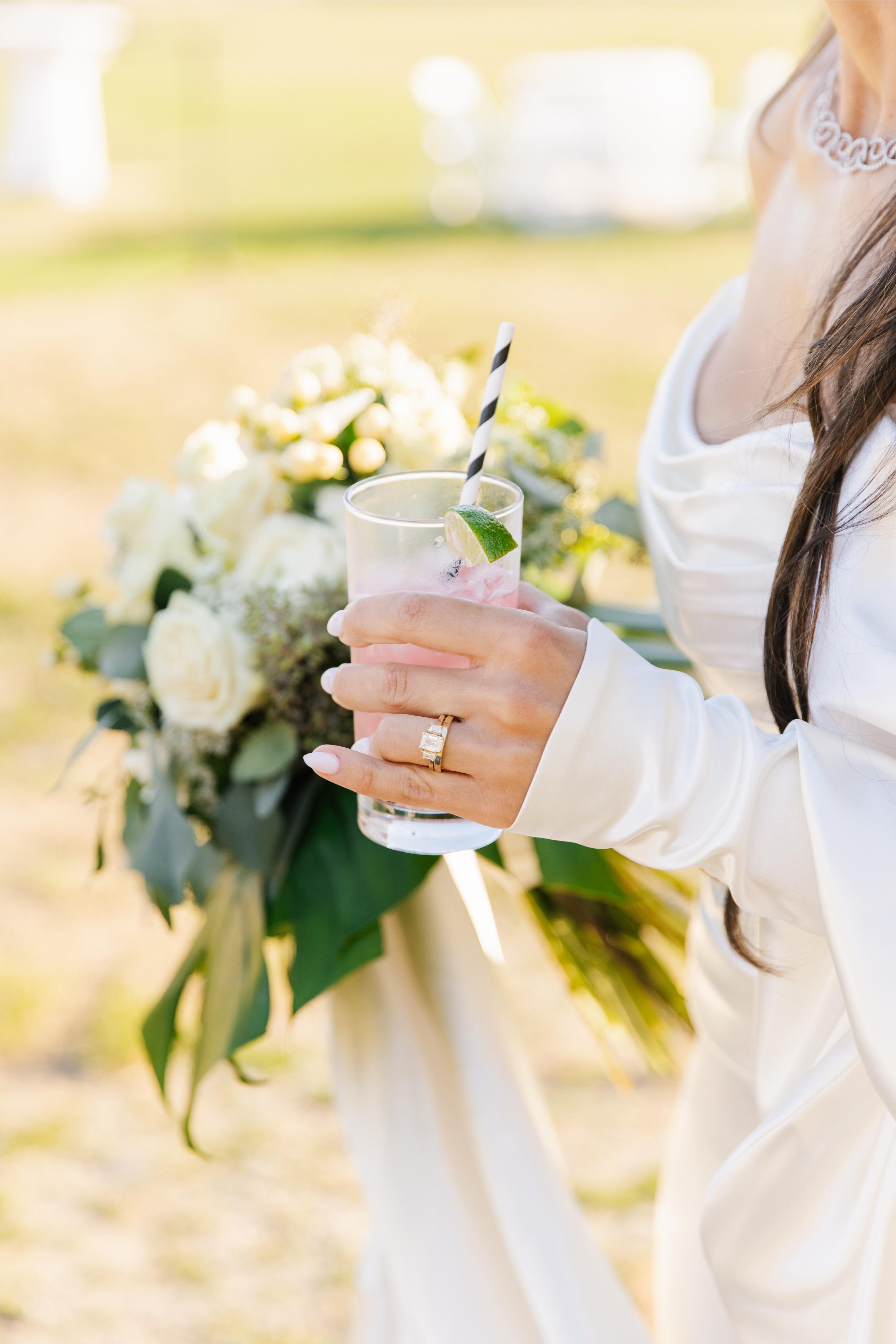 GL Bride Alexandra in Galia Lahav's Alona,  holding a drink with a blurred floral arrangement in the background