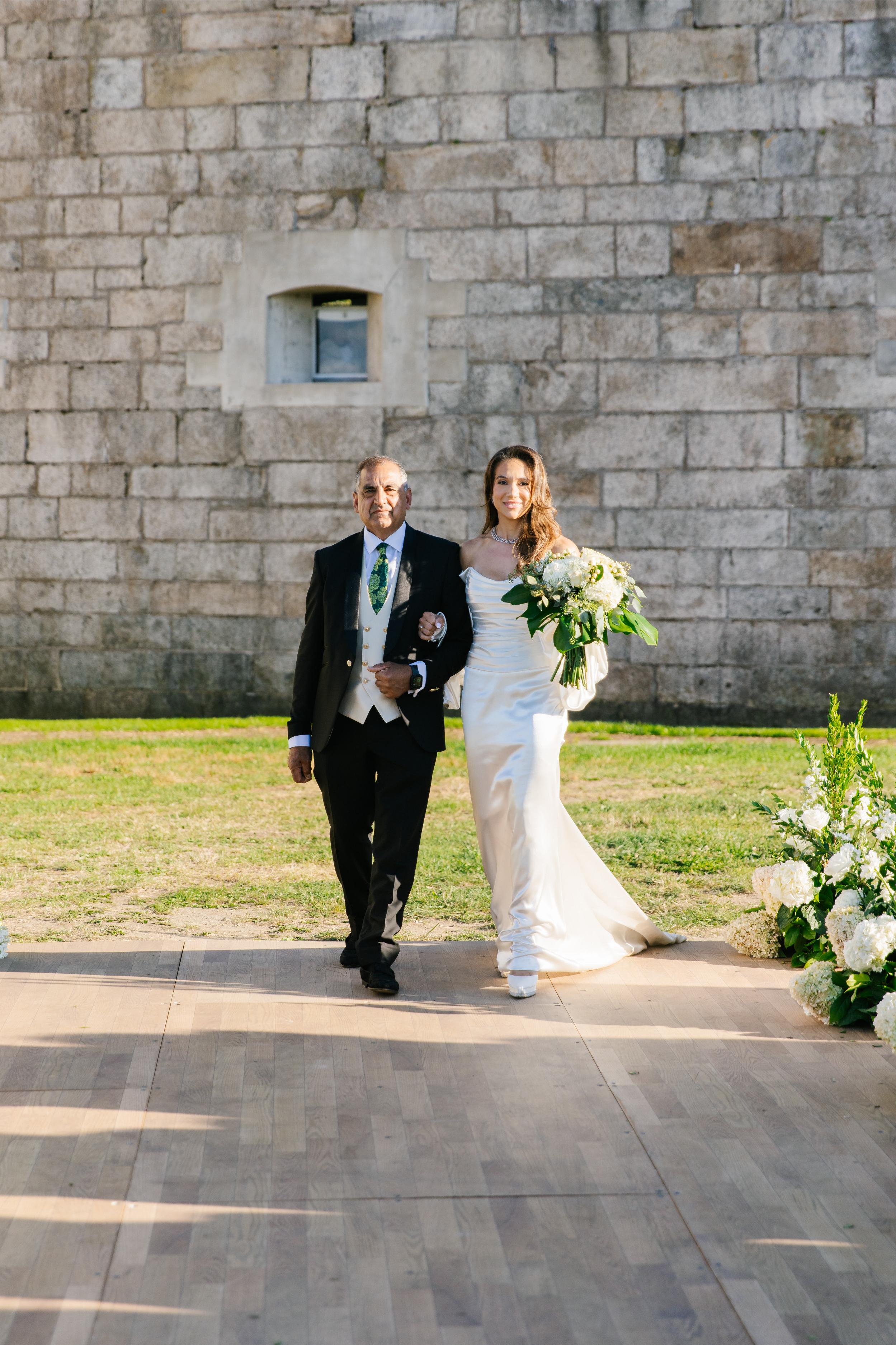 GL Bride Alexandra in Galia Lahav's Alona walking together in front of a stone wall with floral arrangements on either side.