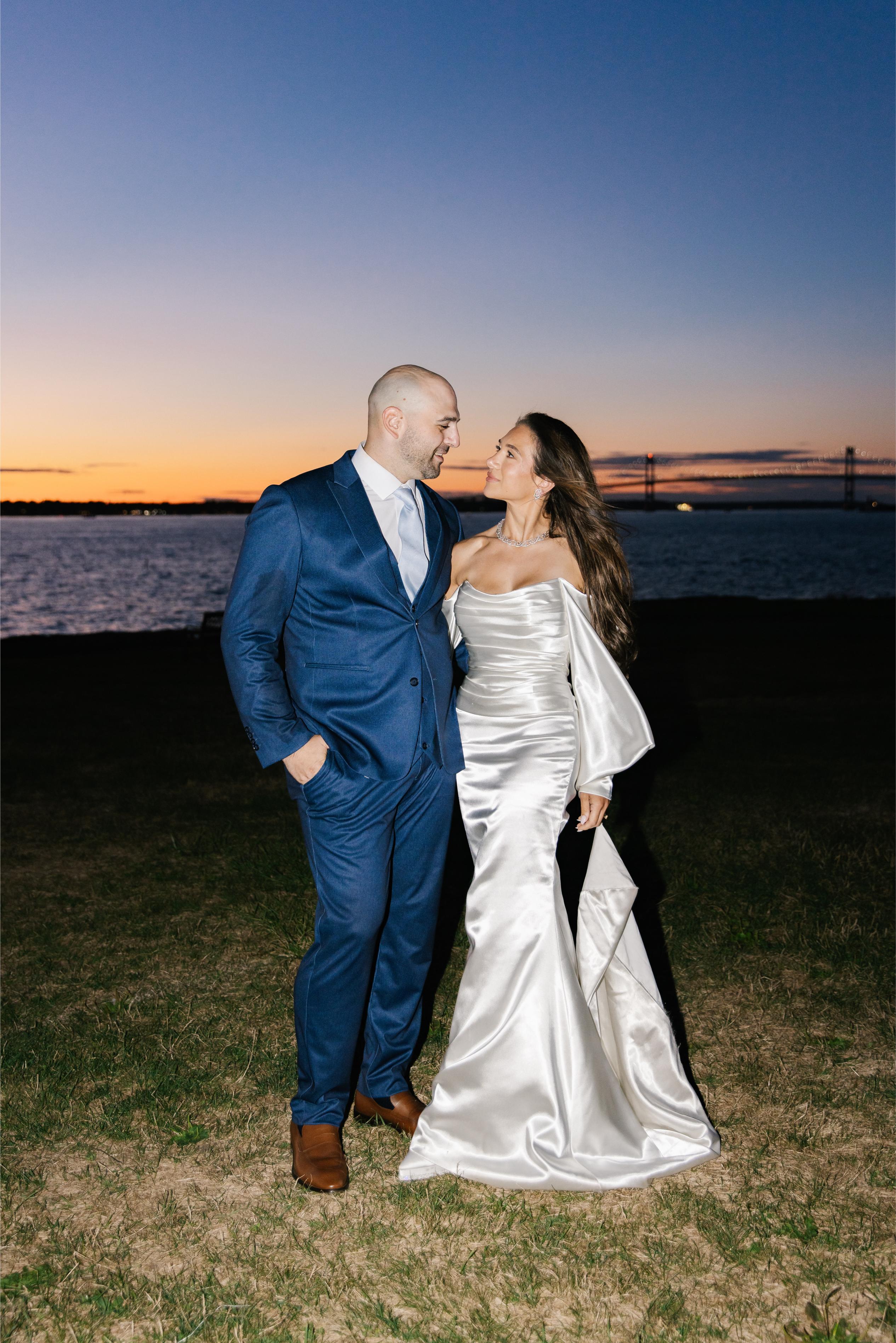 GL Bride Alexandra in Galia Lahav's Alona standing with her husband on the grass with water and sunset in the background