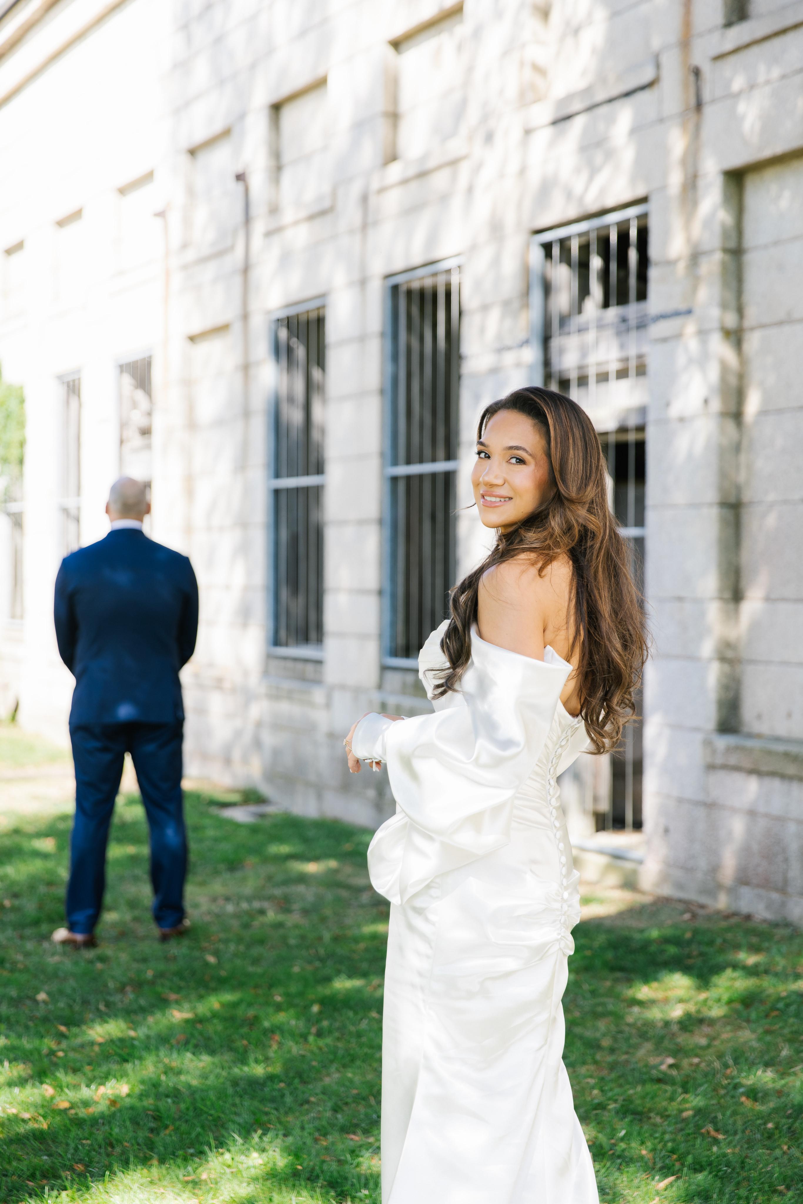 GL Bride Alexandra in Galia Lahav's Alona standing in front of a building with a man in a suit in the background.