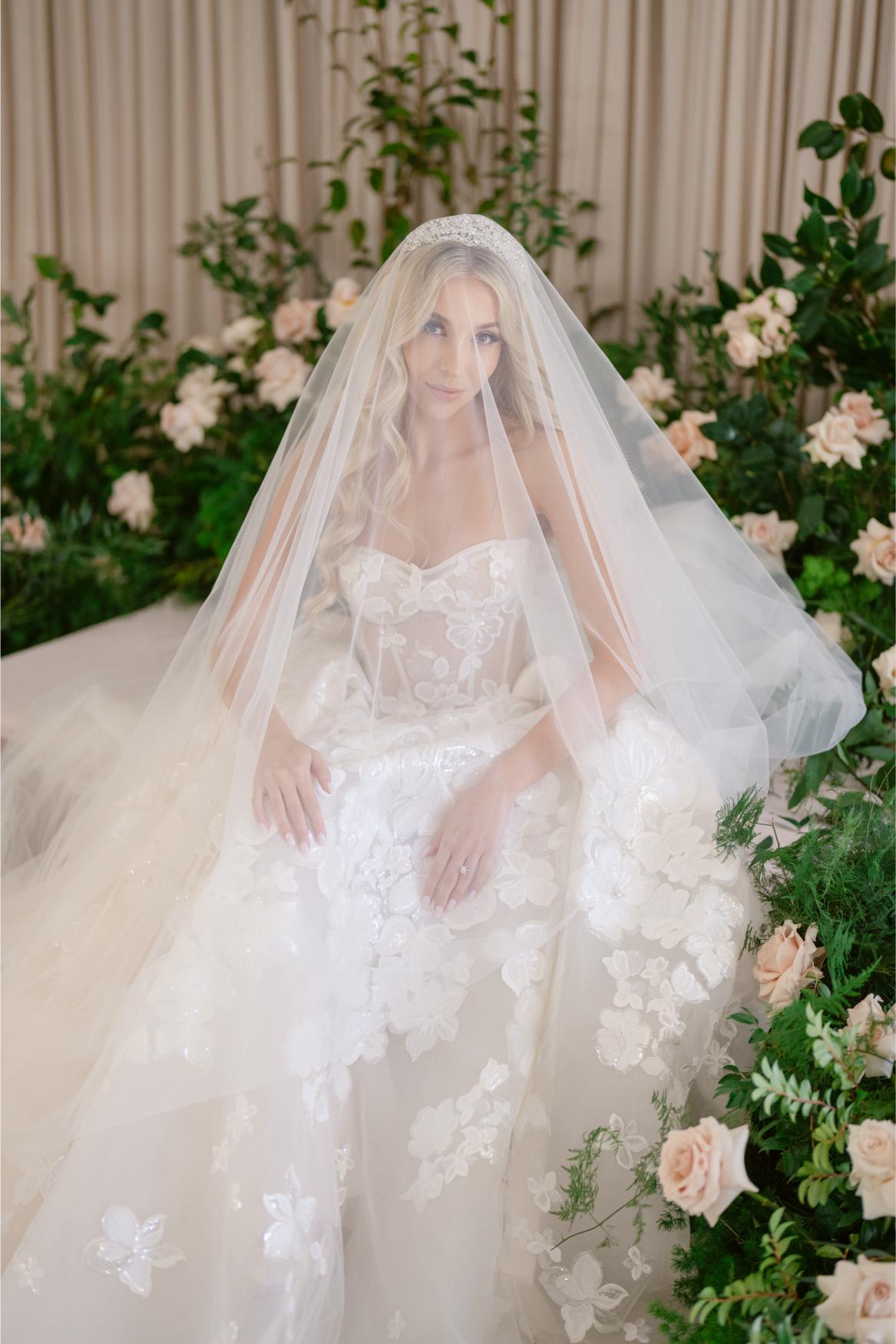 GL Bride Anaies in Renee wedding dress with a veil standing among flowers and greenery.
